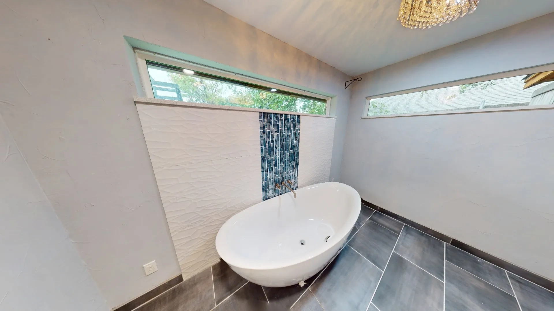 Freestanding soaking tub with wall-mounted plumbing, textured tile accent wall with integrated blue mosaic inlay, large clerestory windows, and large-format porcelain tile flooring