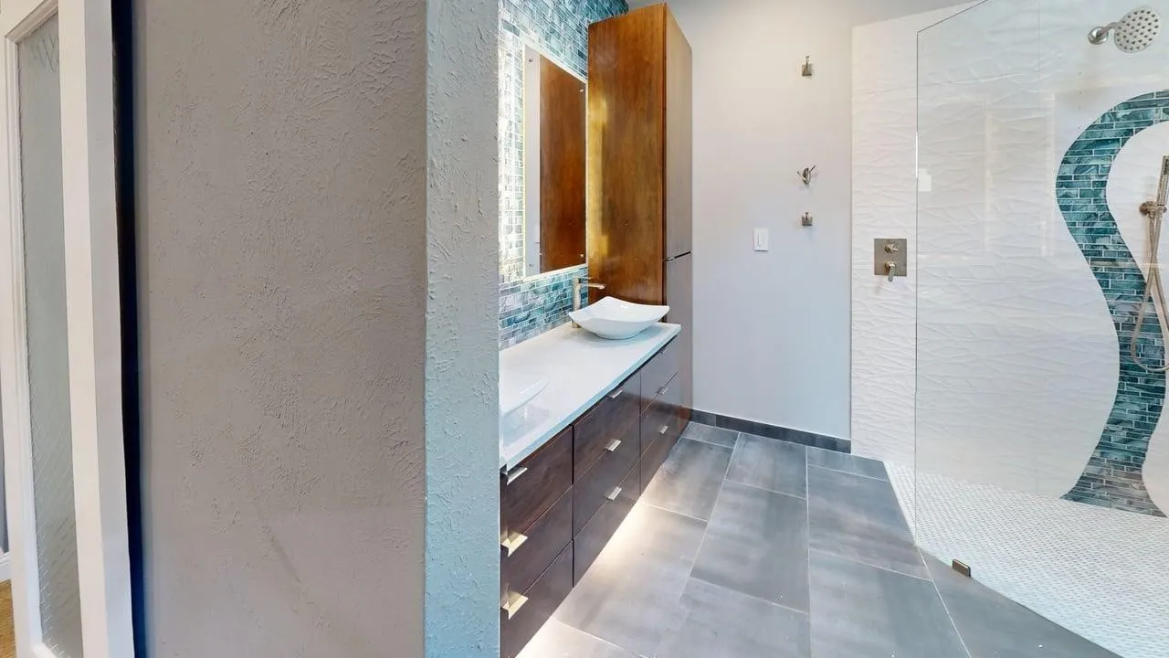 Bathroom featuring wood-finish cabinetry, a white vessel sink, a stone-finish countertop, under-cabinet lighting, and a glass-enclosed shower with mosaic tile accents