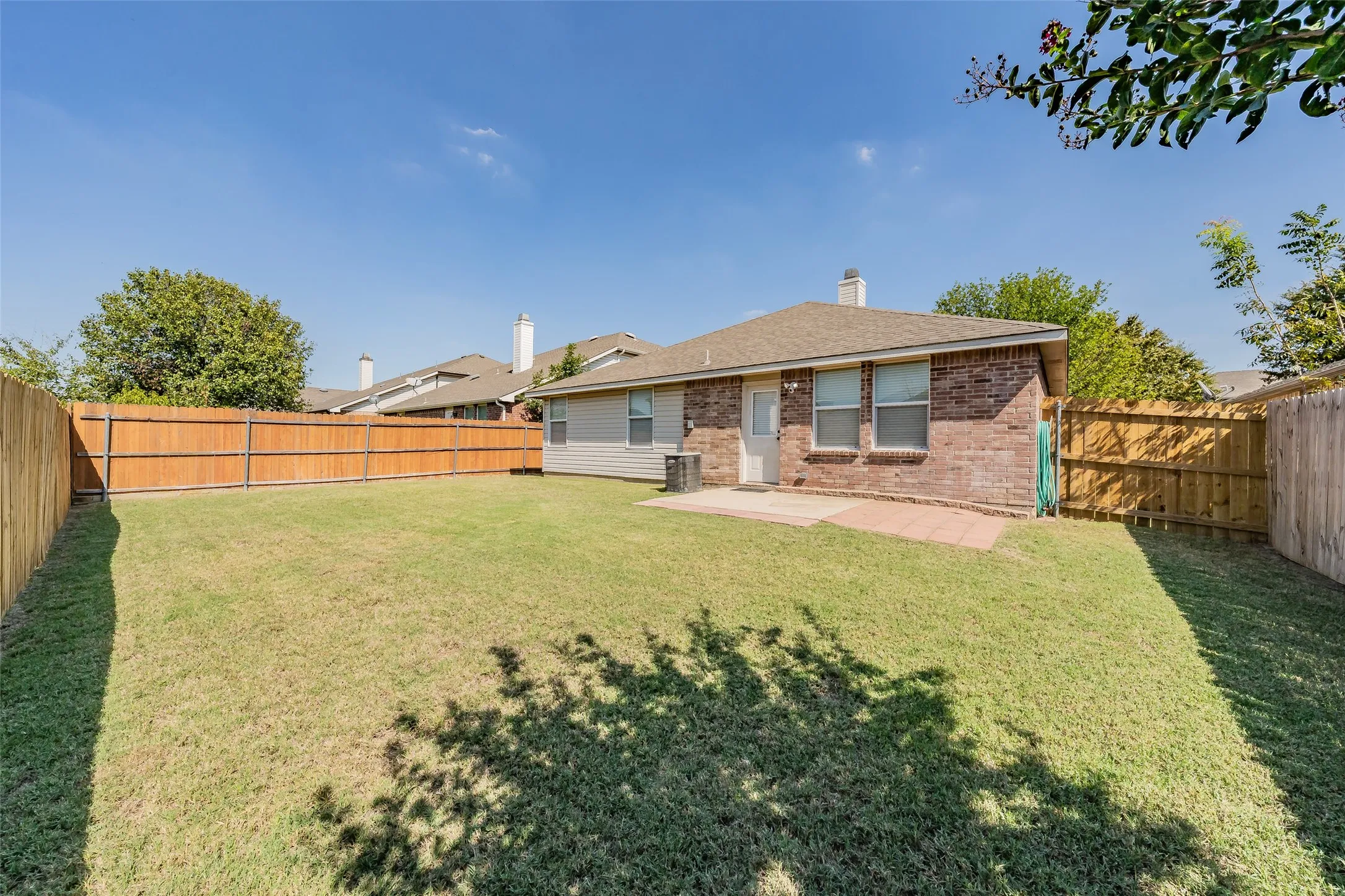 Expansive backyard featuring a manicured lawn, wood fencing, and a brick exterior with an outdoor patio