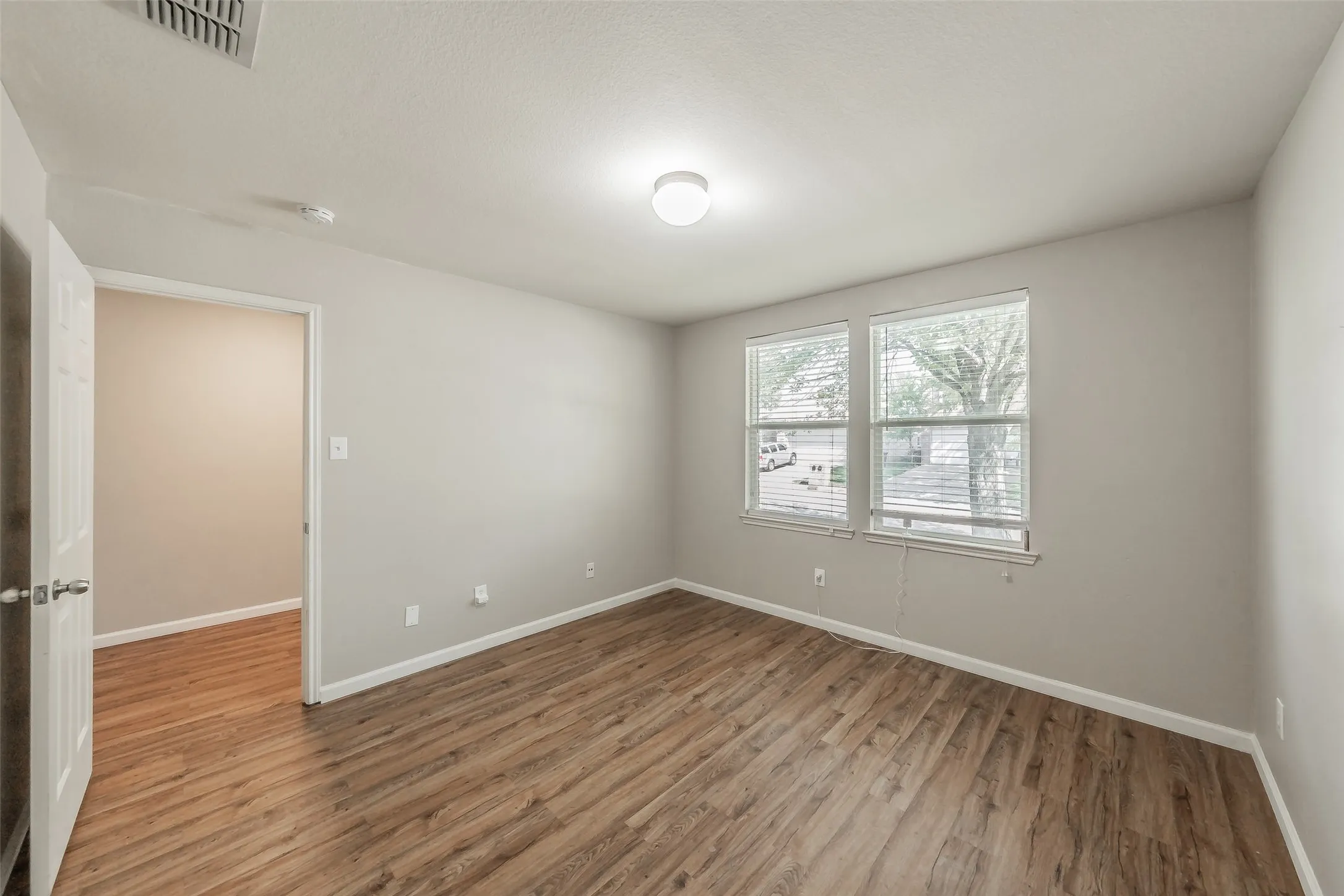 Light gray wall paint, wood-finish flooring, two windows with blinds, white ceiling, and a flush-mount ceiling light fixture