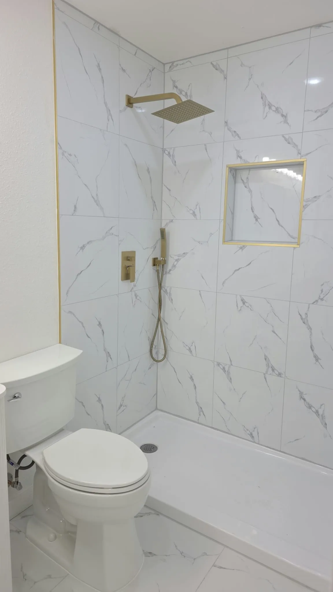 Shower stall featuring white stone-finish tile surround with gold-toned trim, built-in recessed wall niche, rainfall showerhead, and a secondary handheld sprayer with a matching gold-toned finish