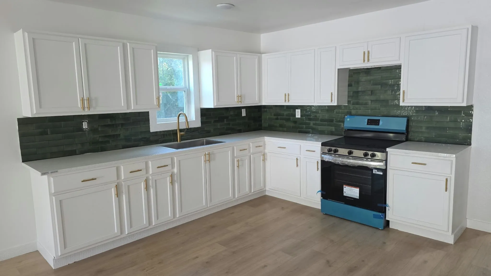 Kitchen featuring white cabinetry with gold-tone hardware, green tile backsplash, light-toned solid surface countertops, and wood-finish flooring