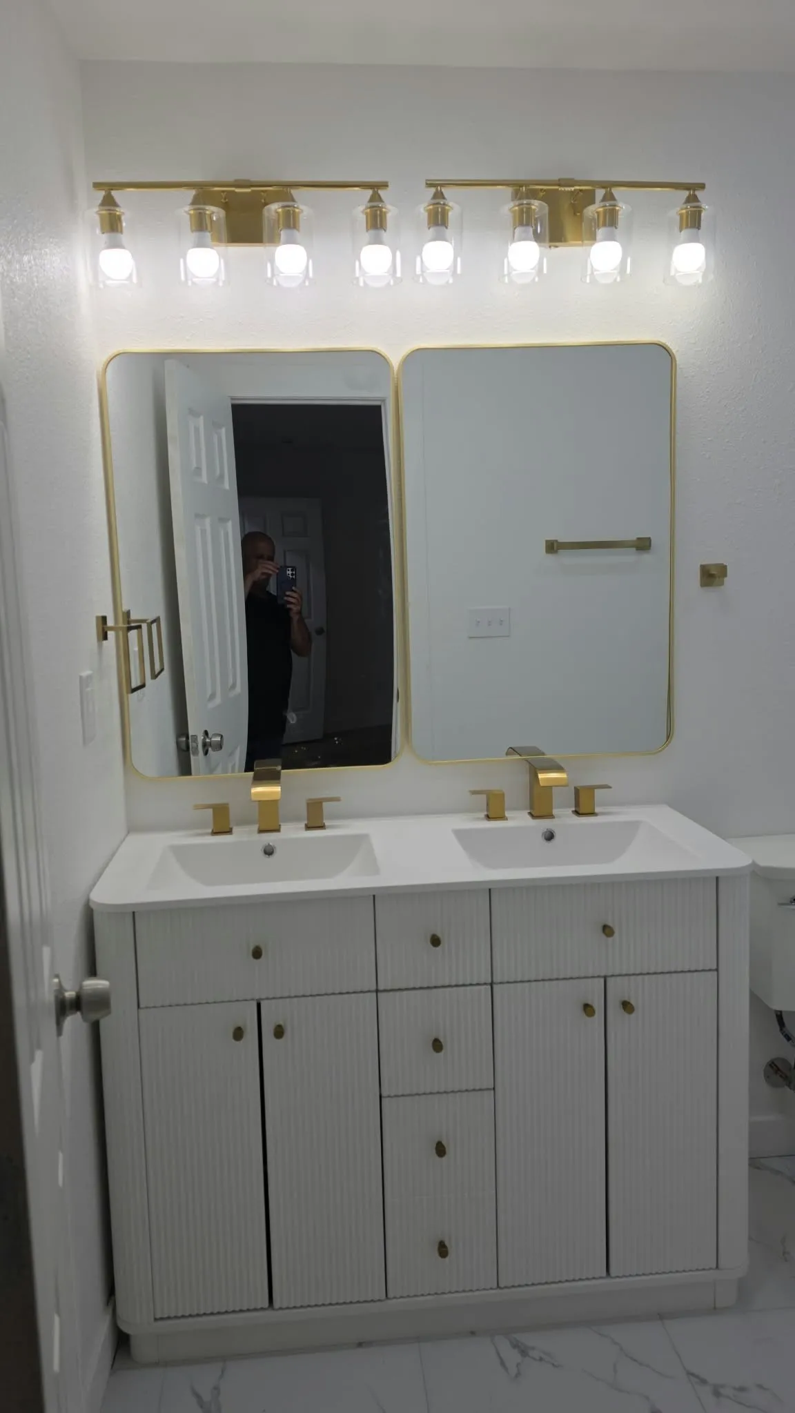 Dual-sink vanity featuring a white fluted cabinet, stone-finish countertop, brass hardware, coordinating dual gold-framed mirrors, and a multi-light brass fixture