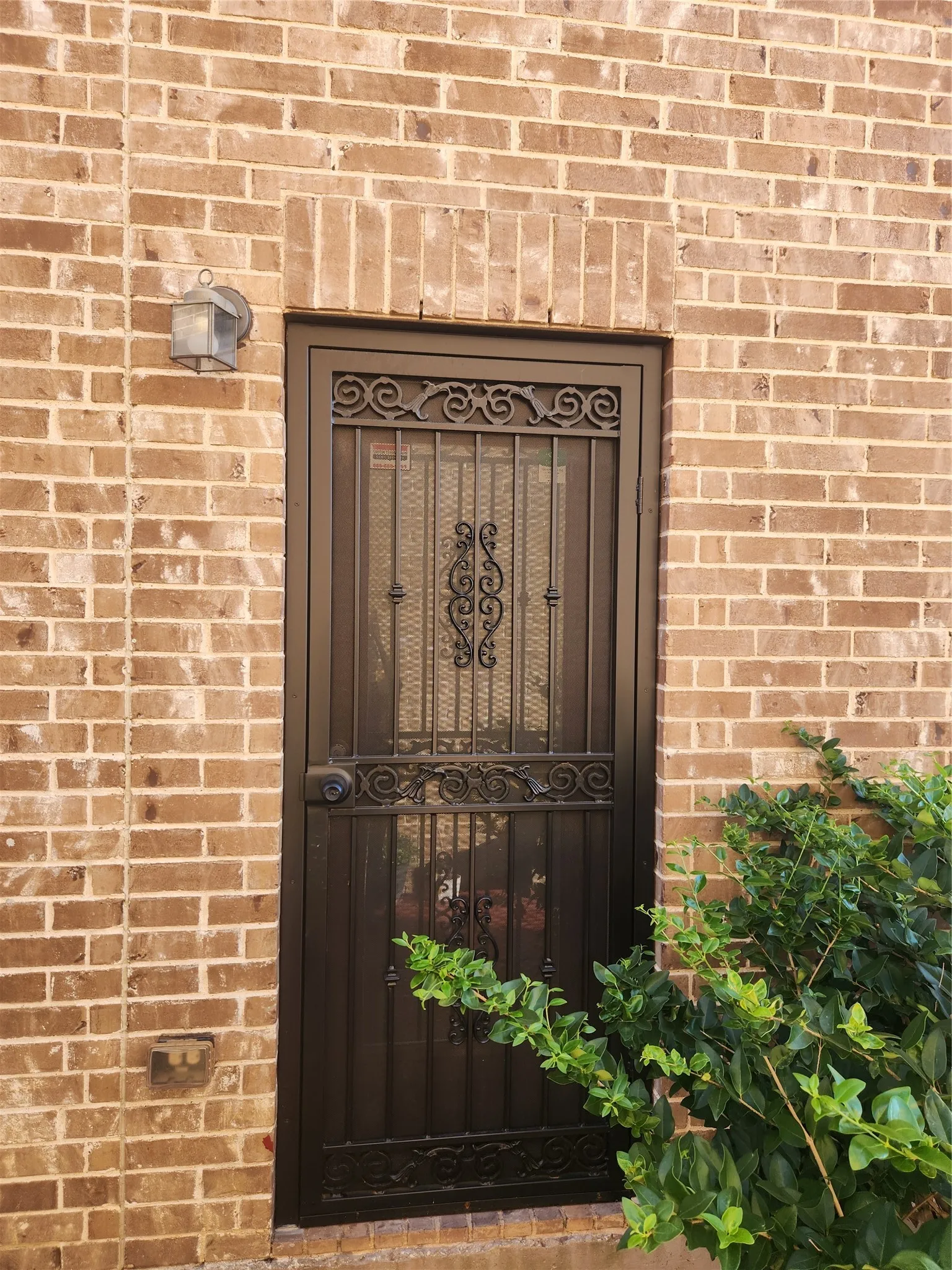 Exterior entryway featuring a masonry brick facade, decorative dark metal security door with intricate scrollwork, and a wall-mounted lantern-style exterior light fixture