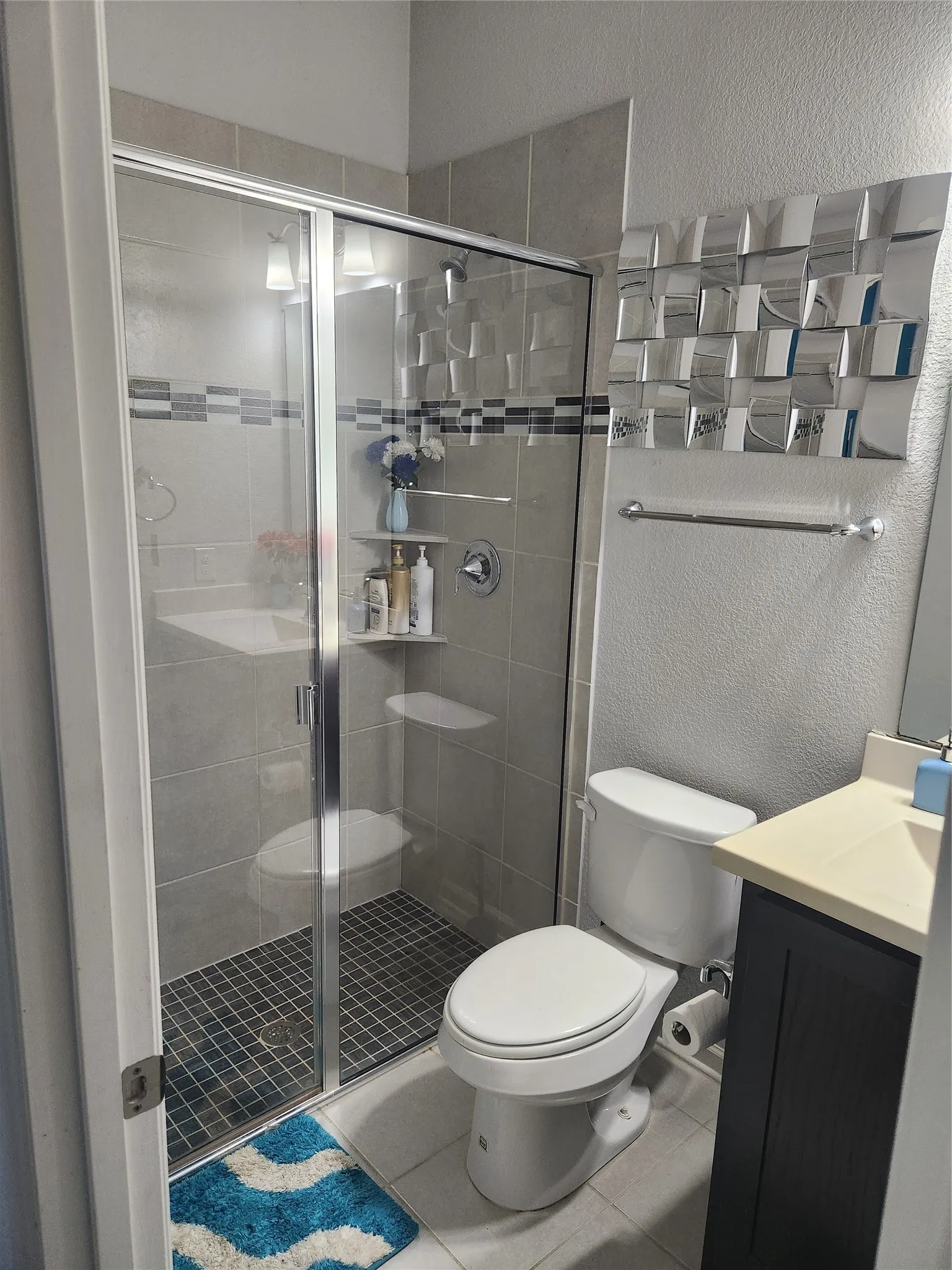 Shower enclosure with glass panels, light gray wall tile, dark gray mosaic shower floor, and an integrated shelf