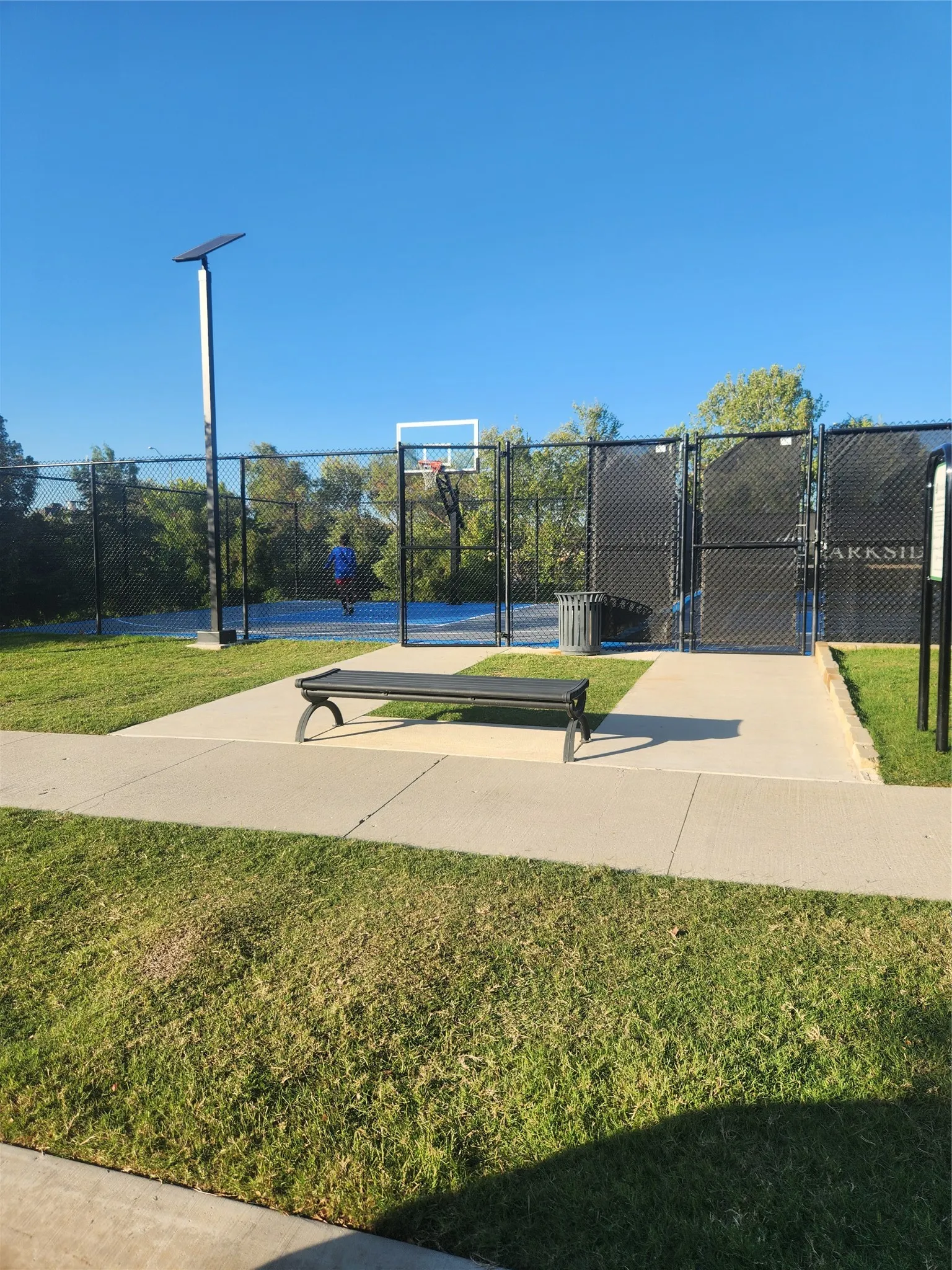 Community basketball court featuring a blue playing surface, black chain-link fencing, and a permanent bench