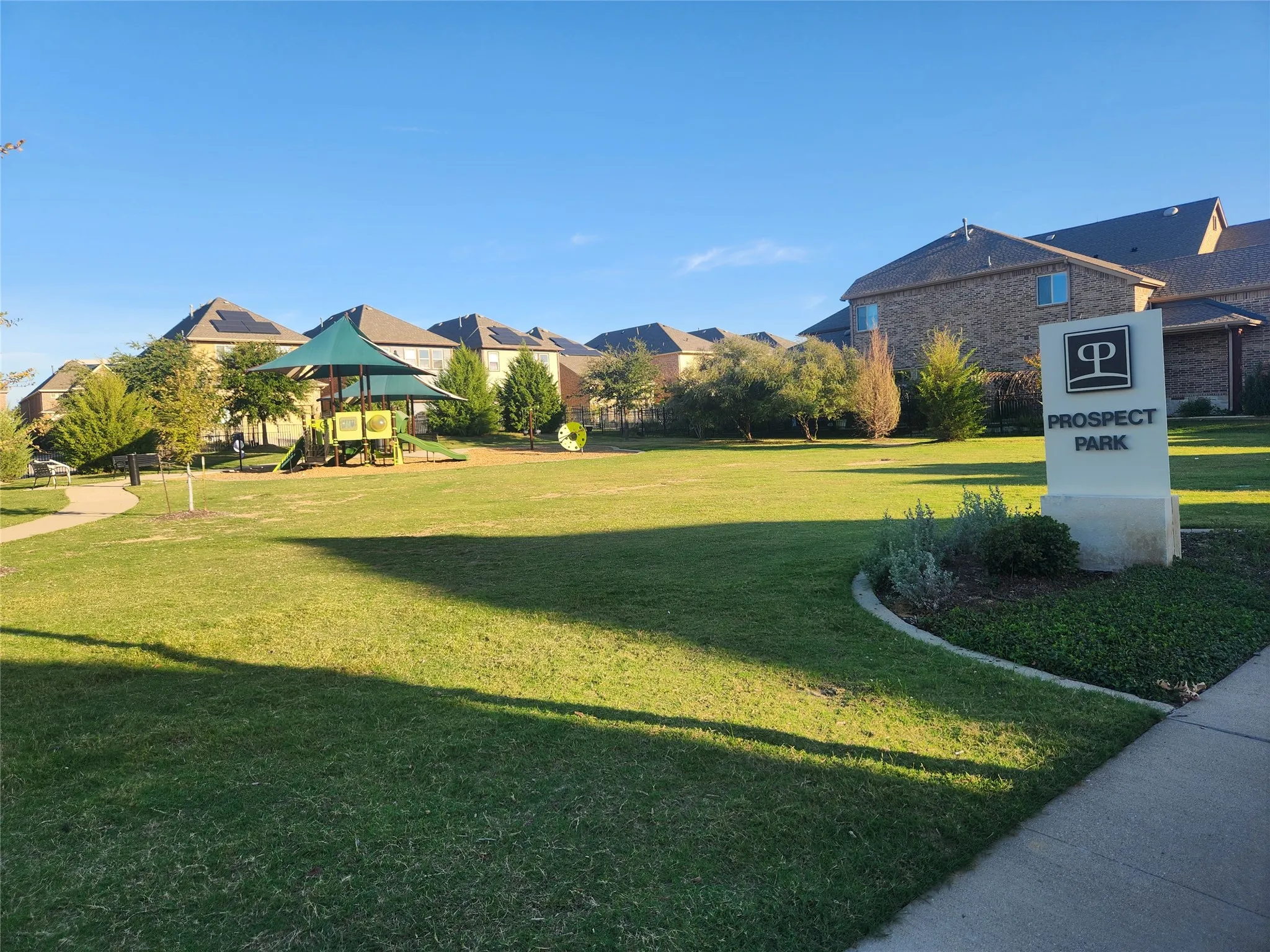 Community park featuring green space, playground structure with shaded canopy, walking paths, and a prominent "Prospect Park" entrance sign