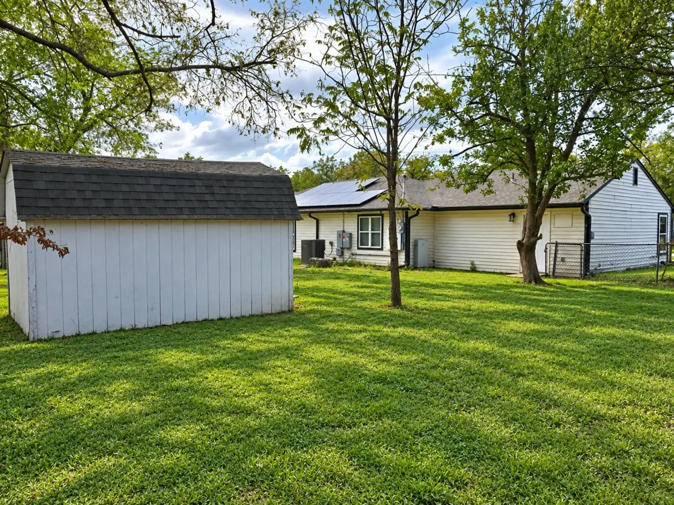 Spacious backyard featuring a detached storage structure, mature trees, chain-link fencing, and a residence equipped with roof-mounted solar panels