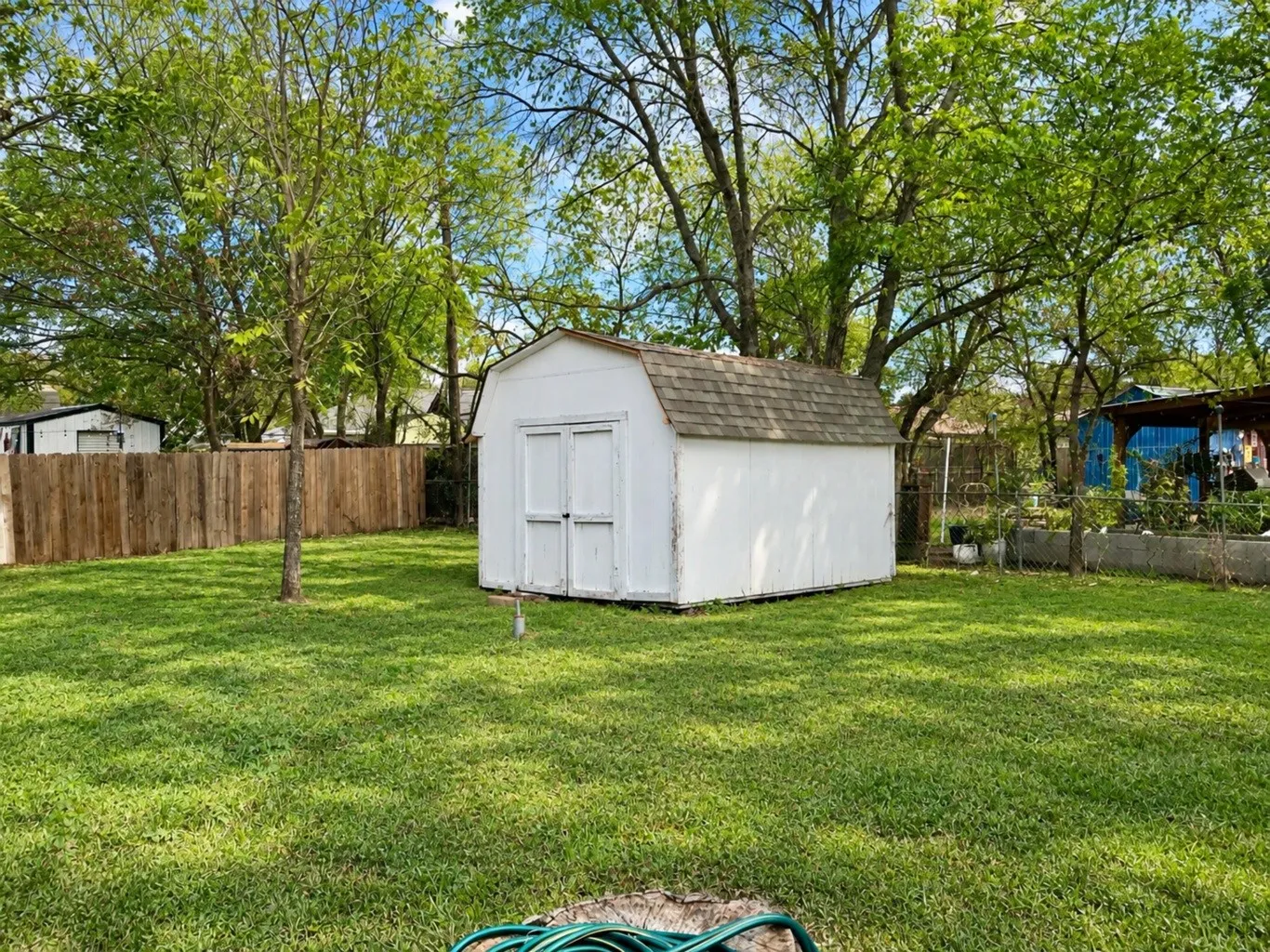 Backyard area featuring a white, gambrel-style storage structure with asphalt shingle roofing, enclosed by a wood privacy fence and established trees