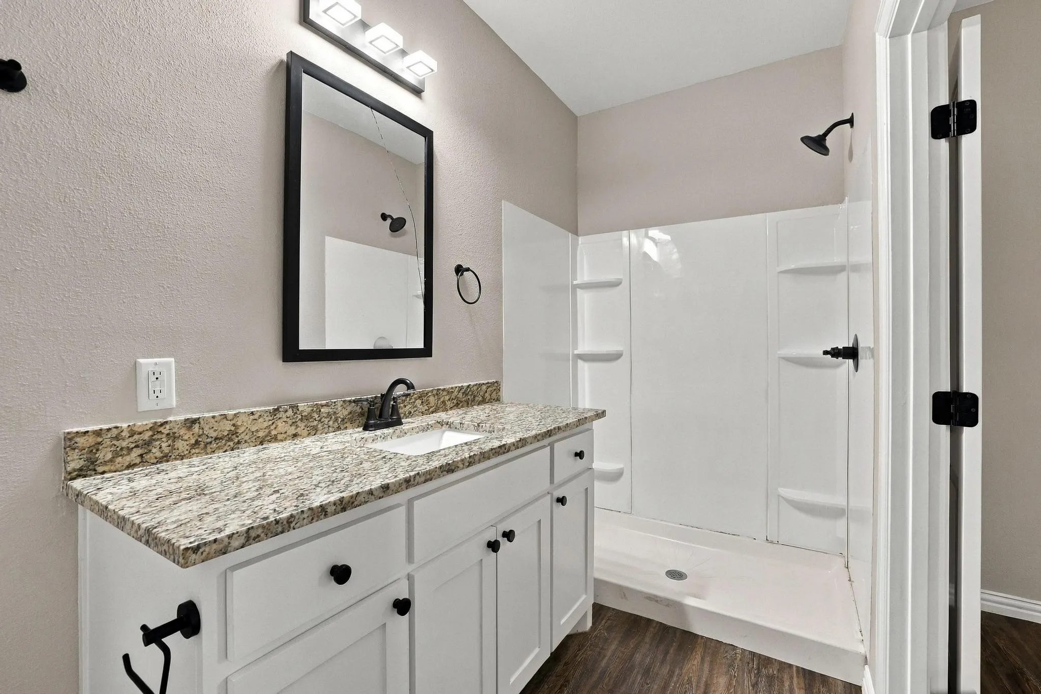 Bathroom featuring a white vanity cabinet, patterned stone countertop with an integrated sink, matte black faucet and hardware, framed mirror, and a walk-in shower surround