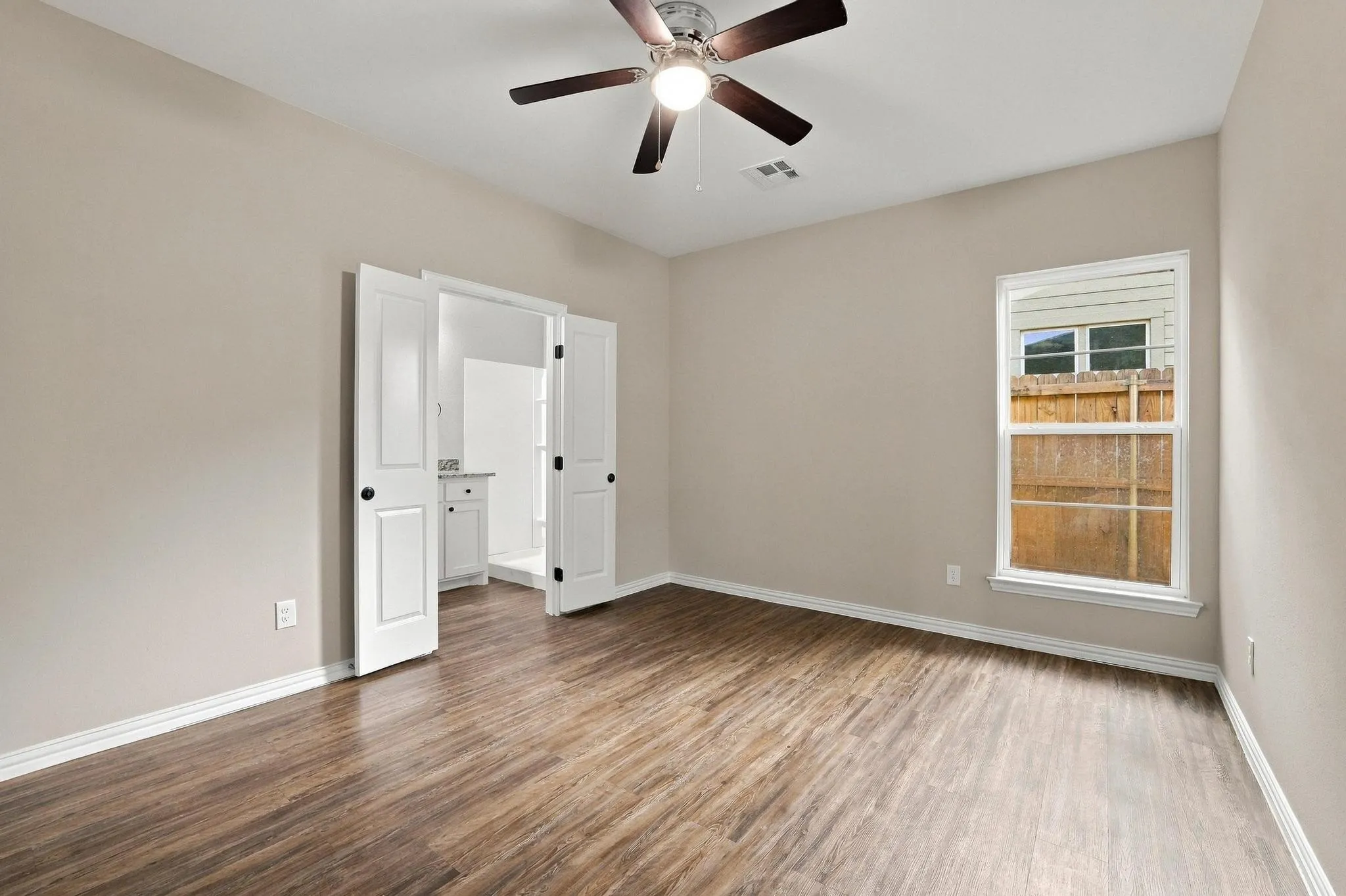 Neutral-toned room featuring wood-finish flooring, a multi-blade ceiling fan with integrated lighting, and dual hinged doors