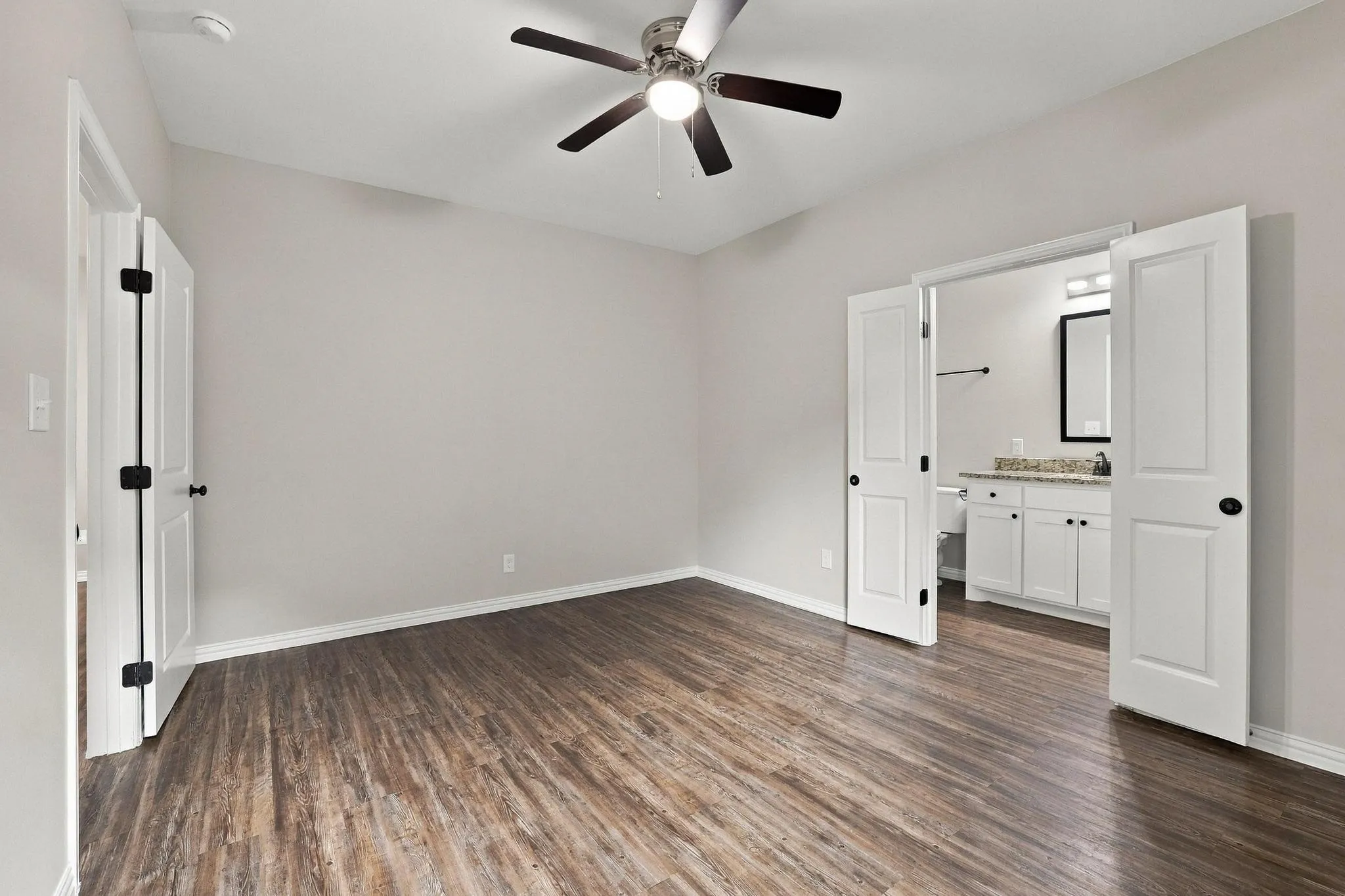 Neutral-toned room featuring wood-finish flooring, a central ceiling fan with integrated lighting, and double doors opening to an auxiliary space with a white vanity and stone-surface countertop