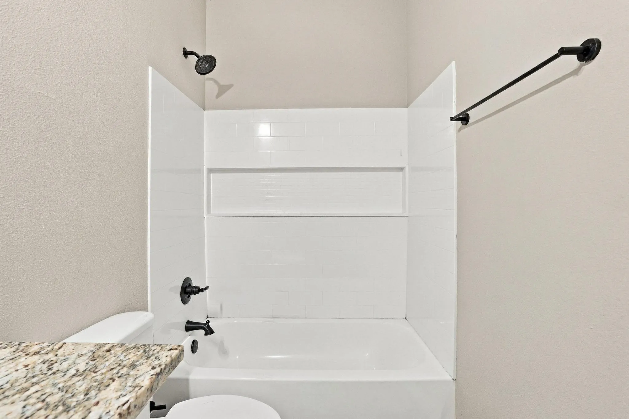 Bathtub and shower combination featuring white surround paneling with integrated recessed shelving, matte black fixtures, and a stone-finish countertop