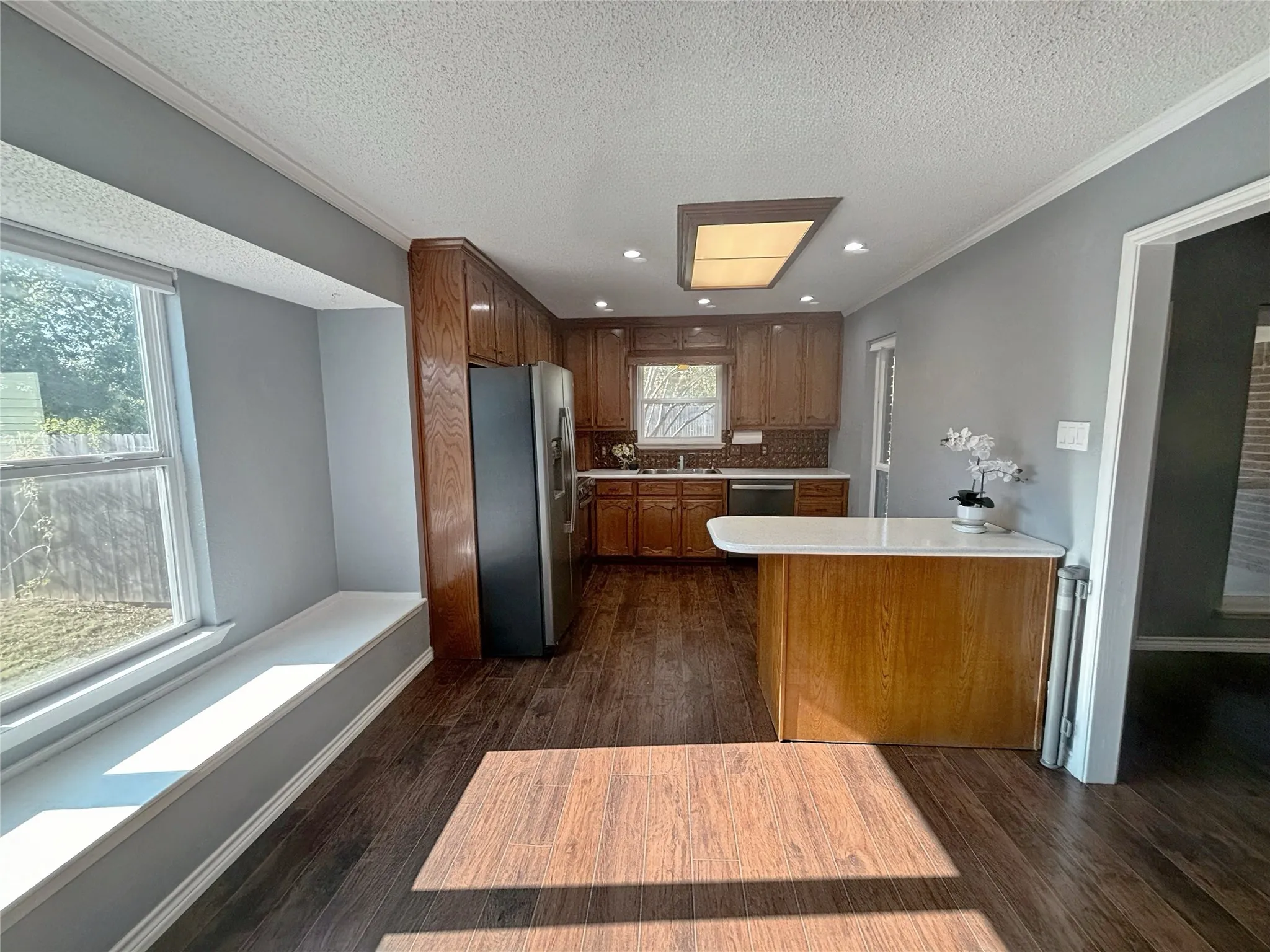 Kitchen featuring wood-finish cabinetry, a peninsula with a stone-surfaced countertop, stainless steel refrigerator, built-in recessed lighting, and a bay window with a structural window seat