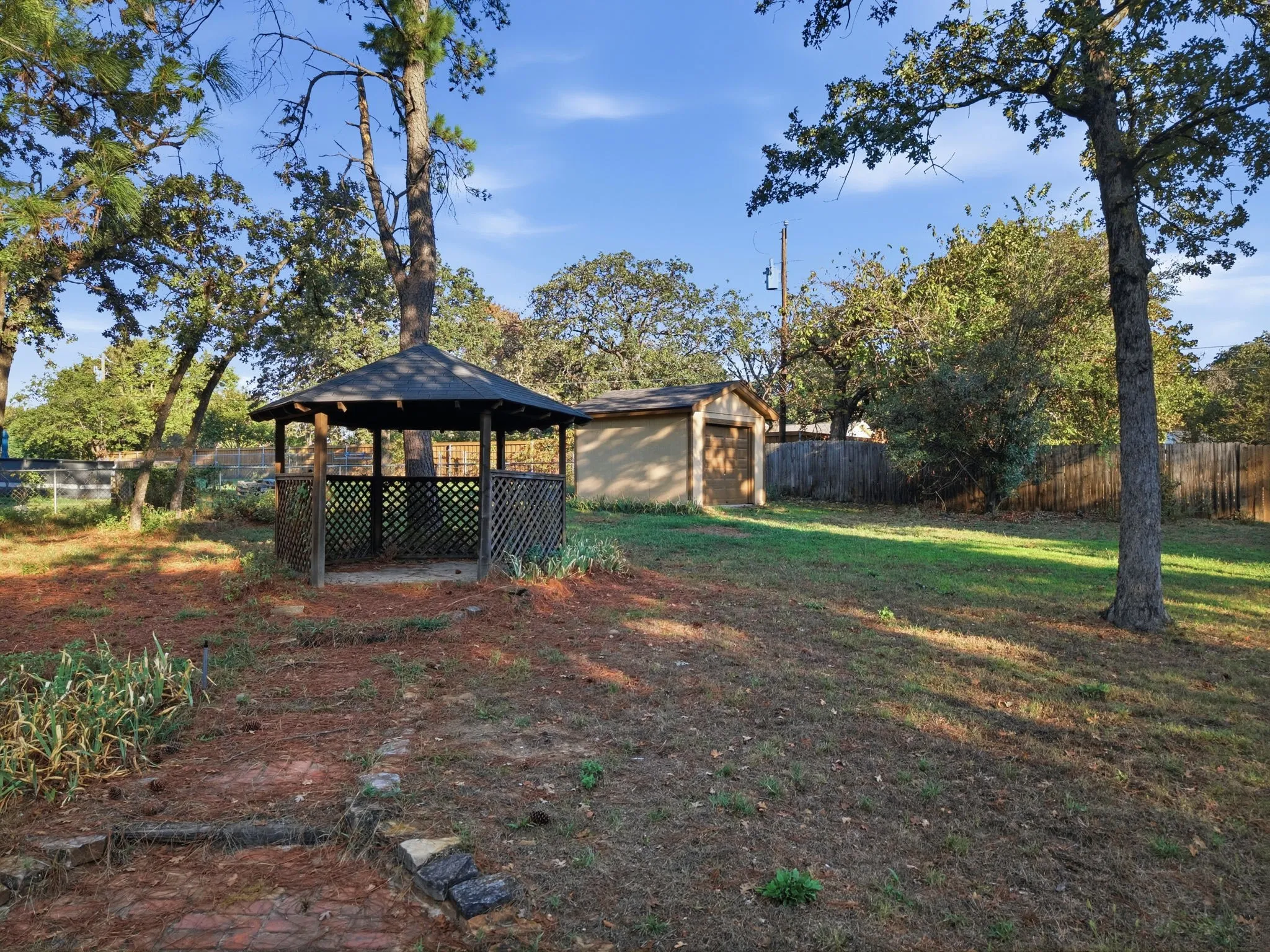 Spacious backyard oasis featuring mature oak trees, a covered gazebo with lattice detailing, a detached storage structure, and lush lawn area ideal for outdoor entertaining and recreation