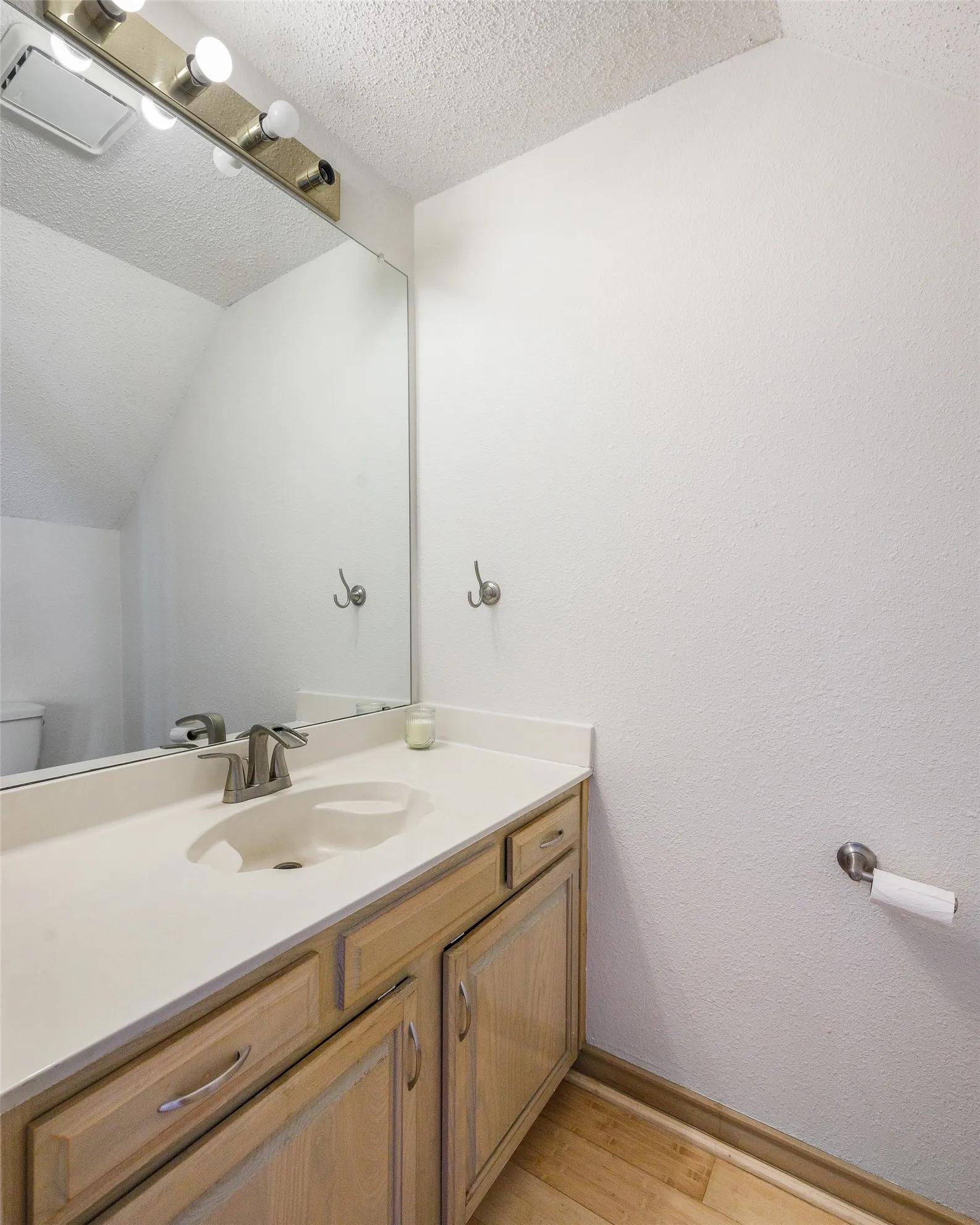 Vanity featuring a light wood-finish cabinet, solid-surface countertop with an integrated basin, wall-to-wall mirror, and multi-bulb fixture