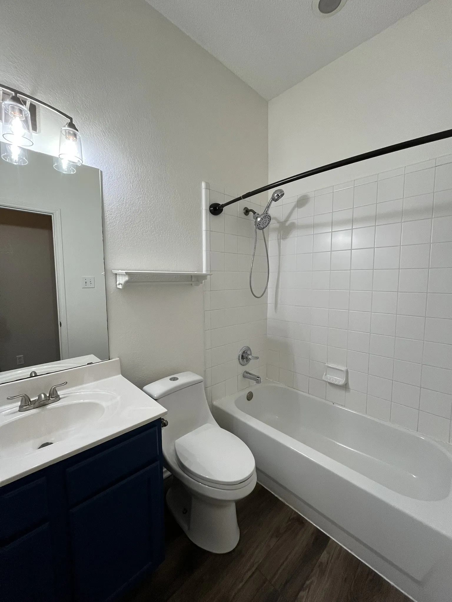 Wood-finish flooring, white subway tile bath surround, dark blue vanity with integrated sink, modern two-bulb vanity light, and a white bathtub