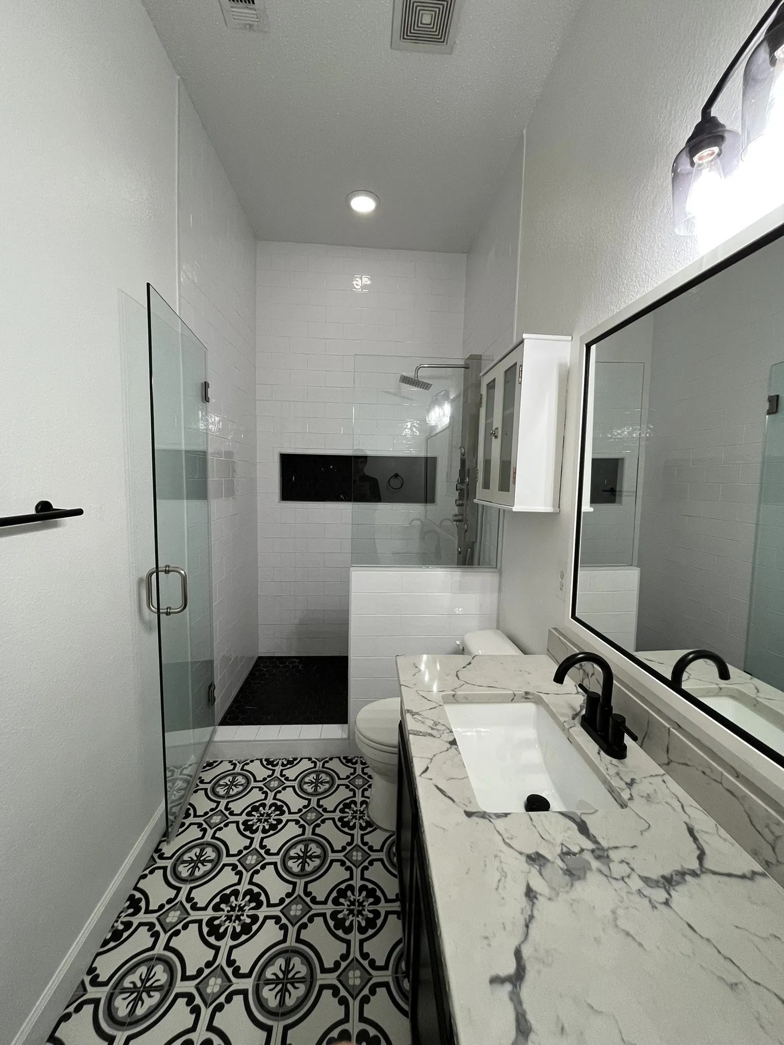 Walk-in shower with glass enclosure, decorative black and white floor tiling, vanity with integral sink and quartz countertop, and black fixtures