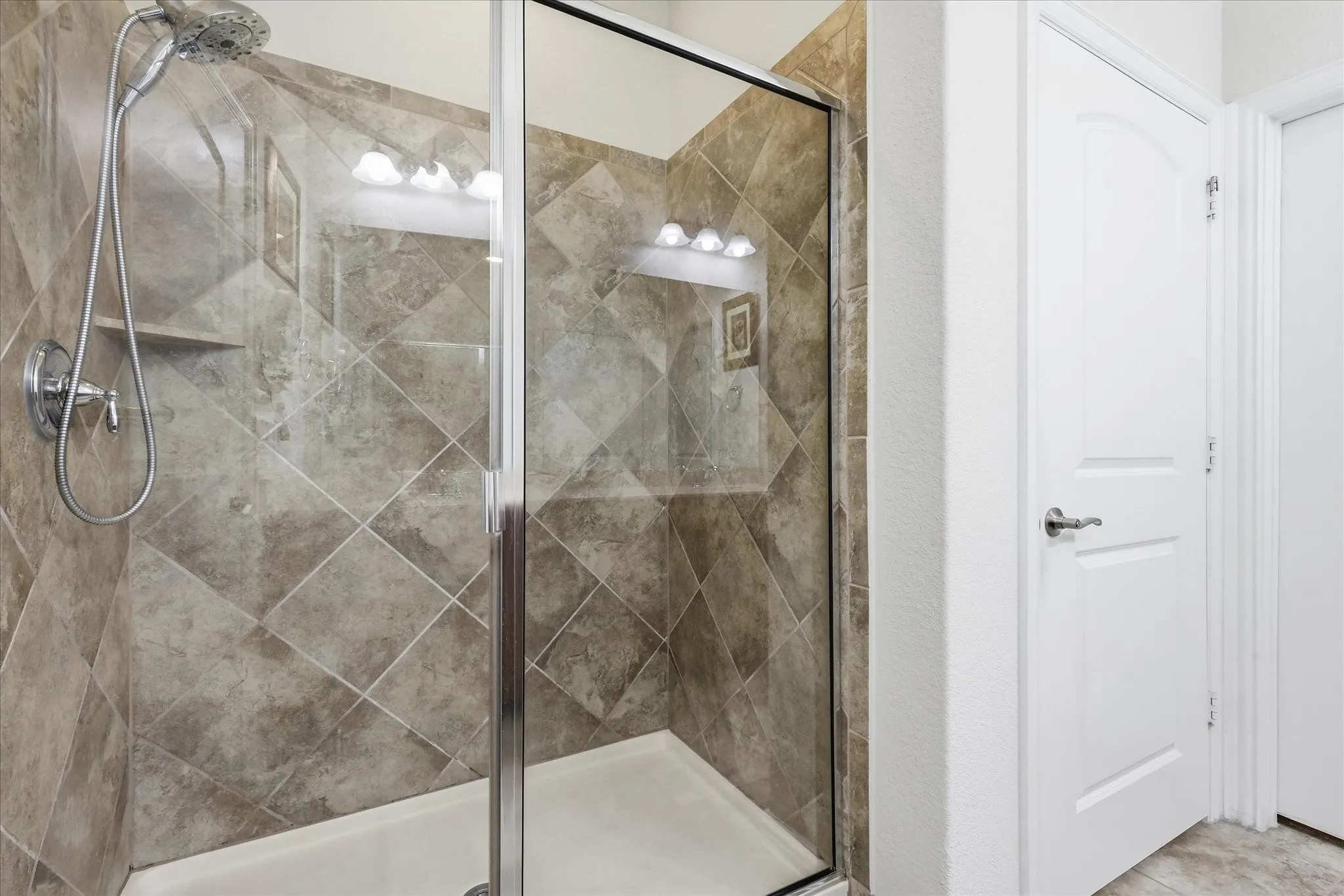 Walk-in shower featuring diamond-patterned tile walls, a handheld showerhead, a built-in recessed shelf, and a glass-enclosed door