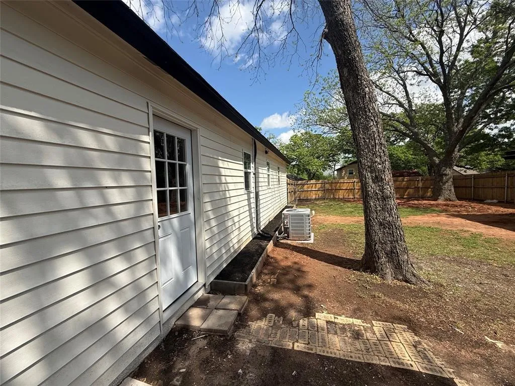 Light-colored horizontal siding exterior with a multi-pane glass door, mature tree, and wooden perimeter fencing