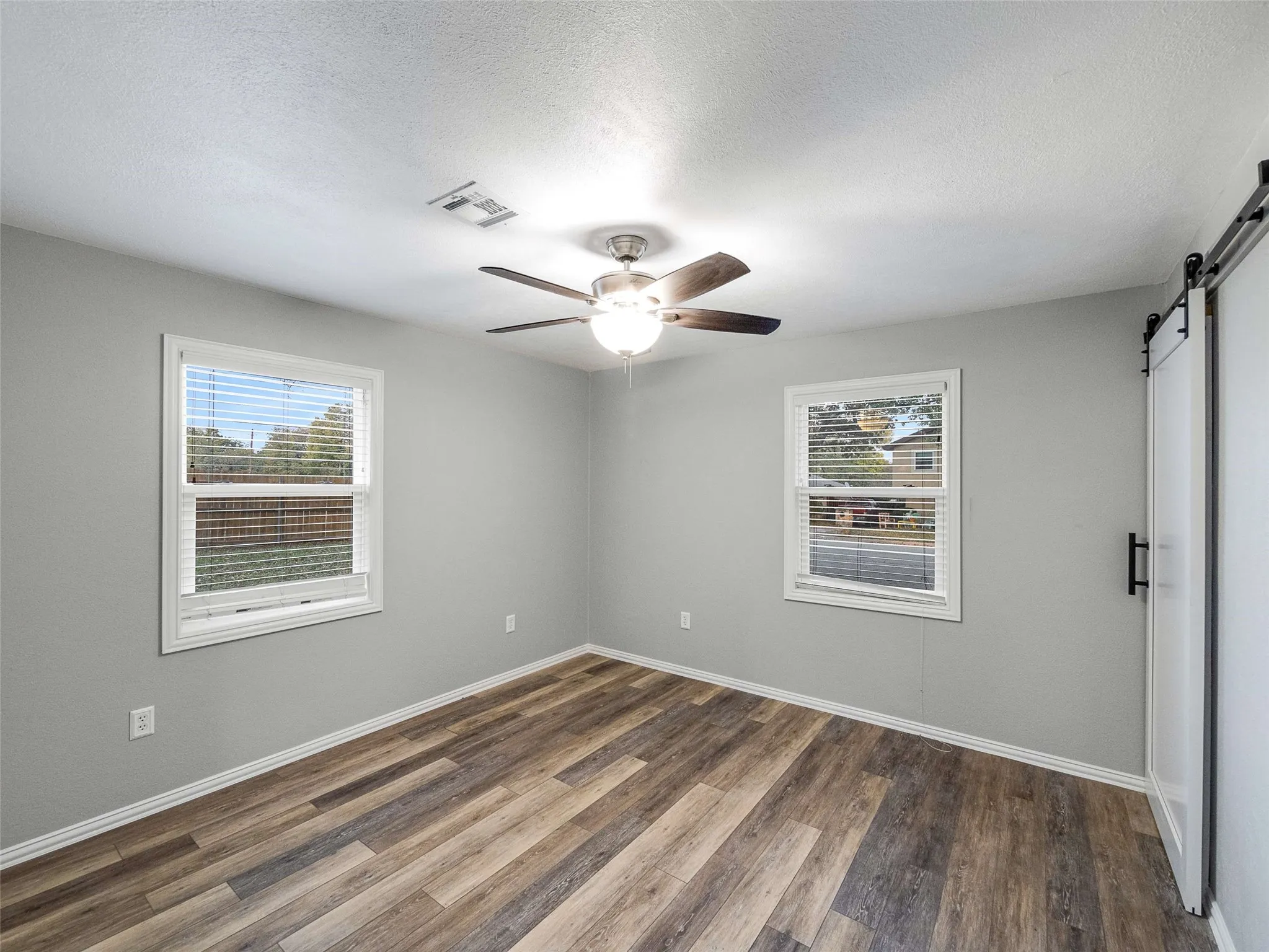 Room featuring wood-finish flooring, dual windows with blinds, a ceiling fan with light fixture, and a white sliding barn door with black hardware