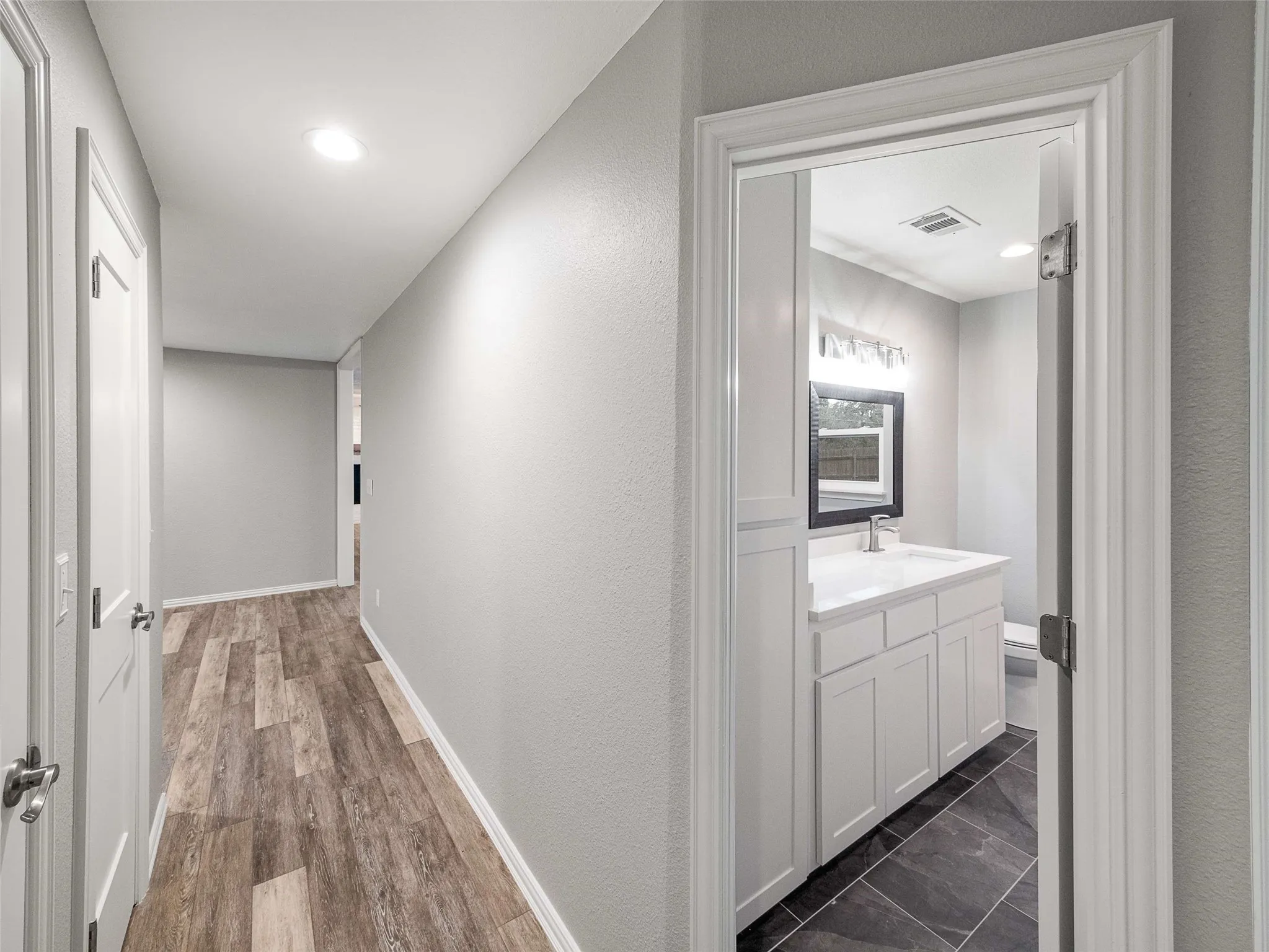 Hallway featuring wood-finish flooring and recessed lighting, opening into a bathroom with a white vanity cabinet, rectangular countertop sink, and large-format tile flooring
