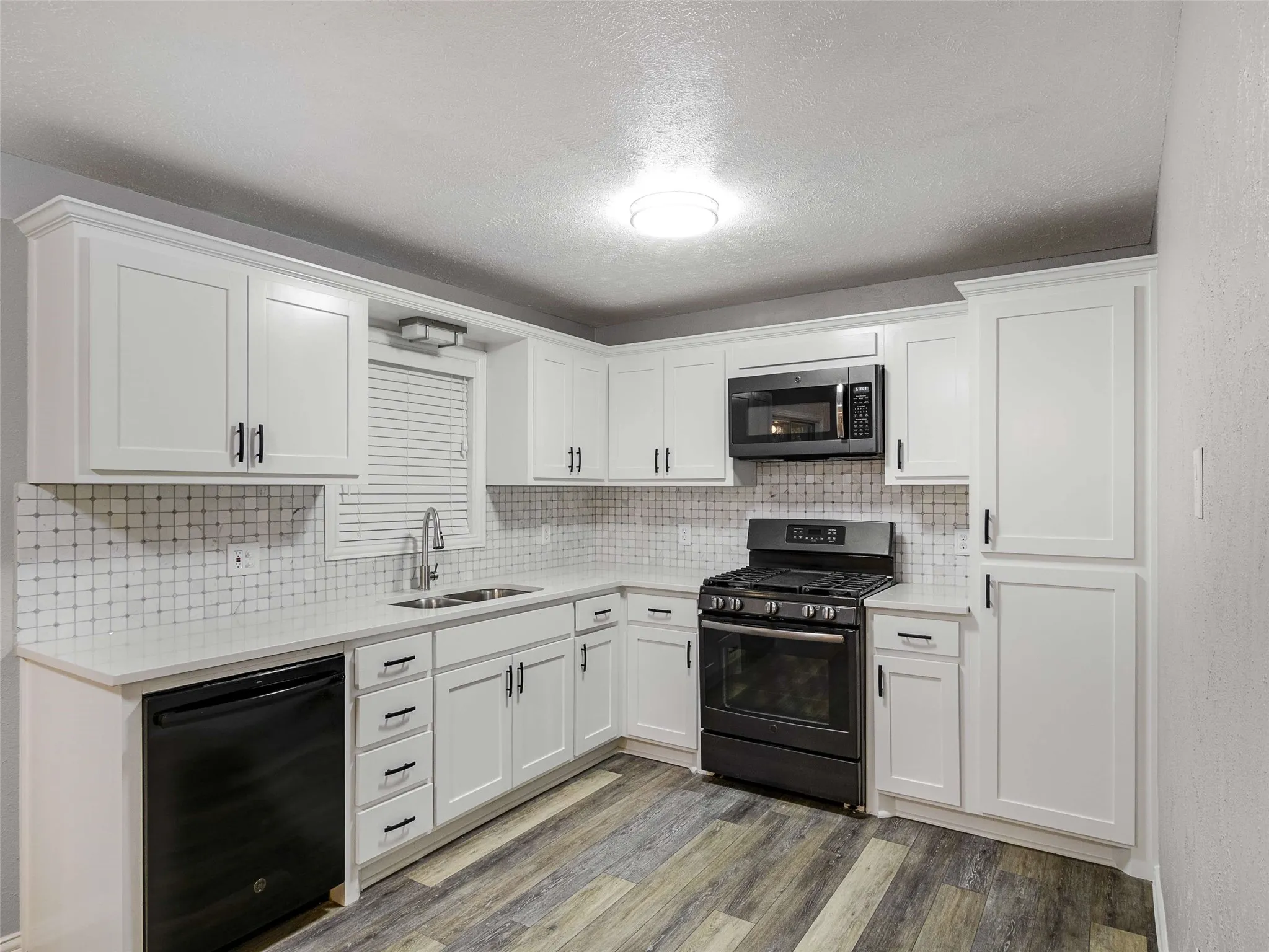 Kitchen featuring white shaker-style cabinetry, light-toned stone-surface countertops, a mosaic tile backsplash, black built-in appliances, and wood-finish flooring