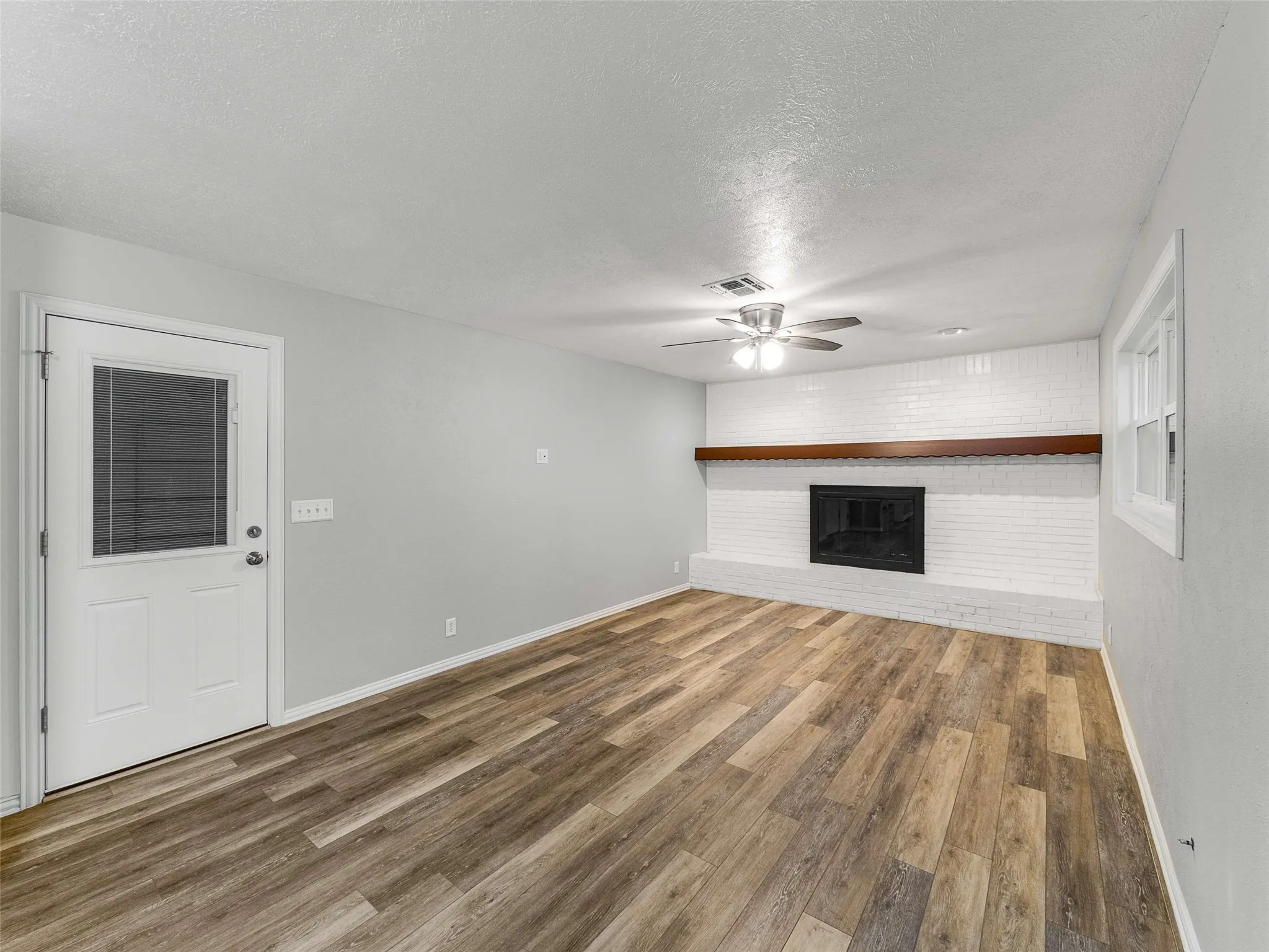 Living area featuring wood-finish flooring, a white-painted brick fireplace, a prominent wooden mantel, and a ceiling fan with integrated lighting