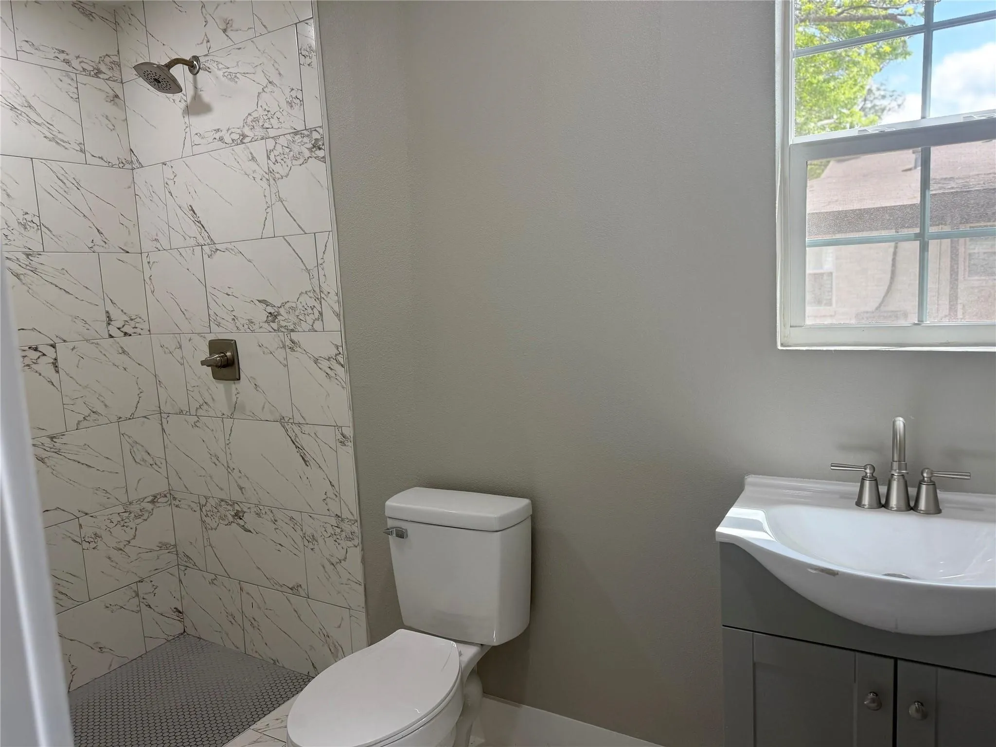Shower stall with patterned tile surround and adjustable wall-mounted shower head, grey vanity cabinet with integrated basin, polished metal faucet, and multi-pane window