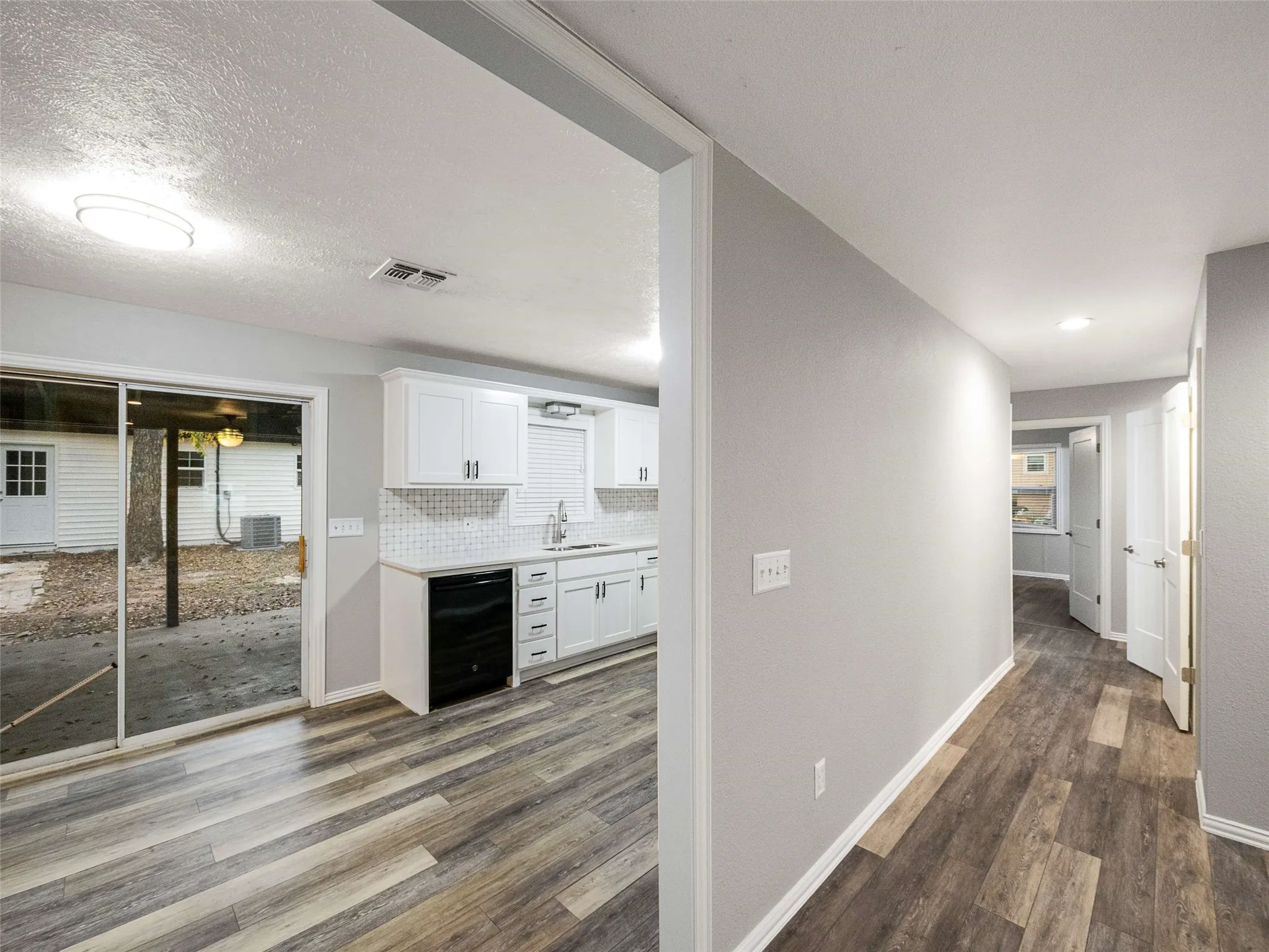 Kitchen featuring white shaker-style cabinetry, mosaic tile backsplash, and a black built-in dishwasher, complemented by wood-finish flooring and a sliding glass door