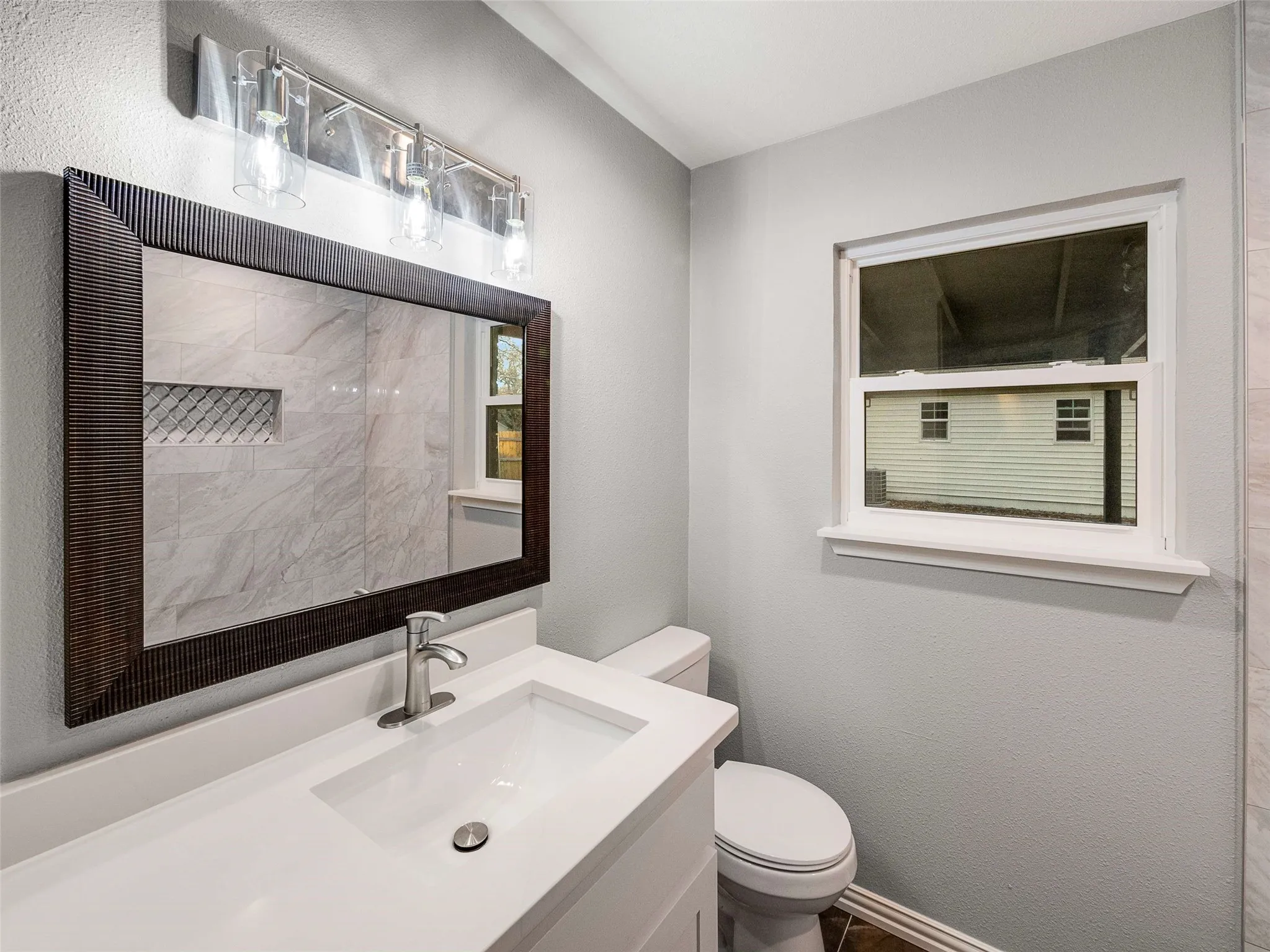 Bathroom vanity featuring a rectangular integrated sink, neutral stone-surface countertop, three-light glass fixture, and framed wall mirror