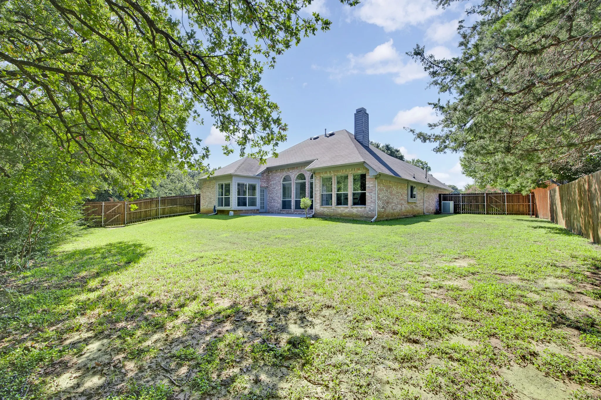 Brick exterior residence featuring a rear-facing chimney, arched windows, expansive lawn, and enclosed wood perimeter fencing