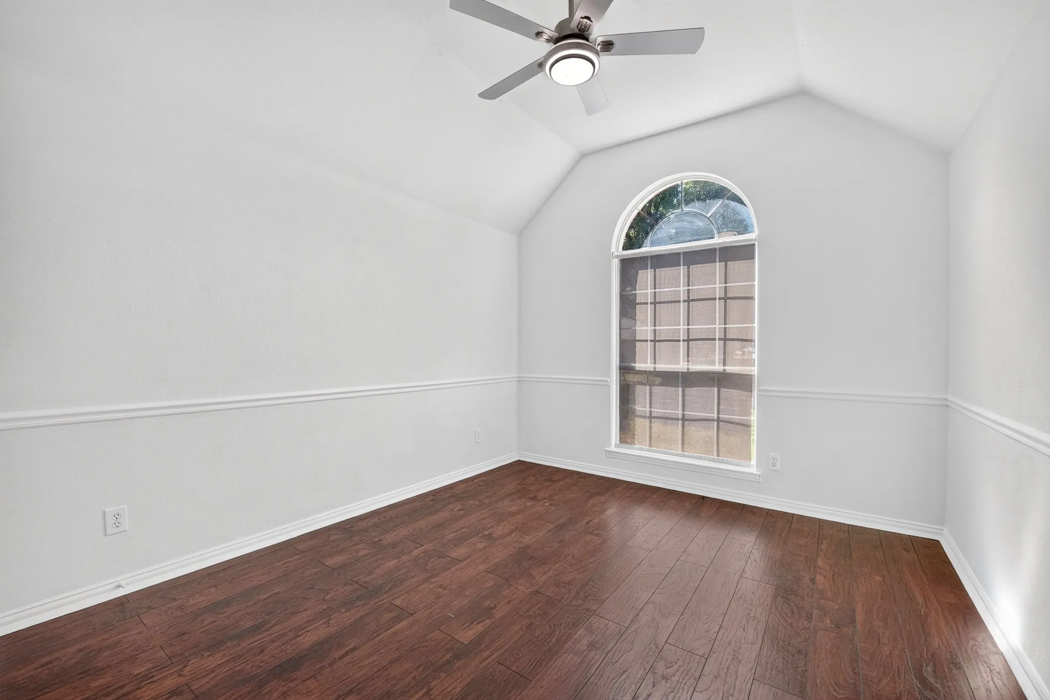Room featuring vaulted ceilings, wood-finish flooring, chair rail molding, an arched window, and a ceiling fan with integrated lighting