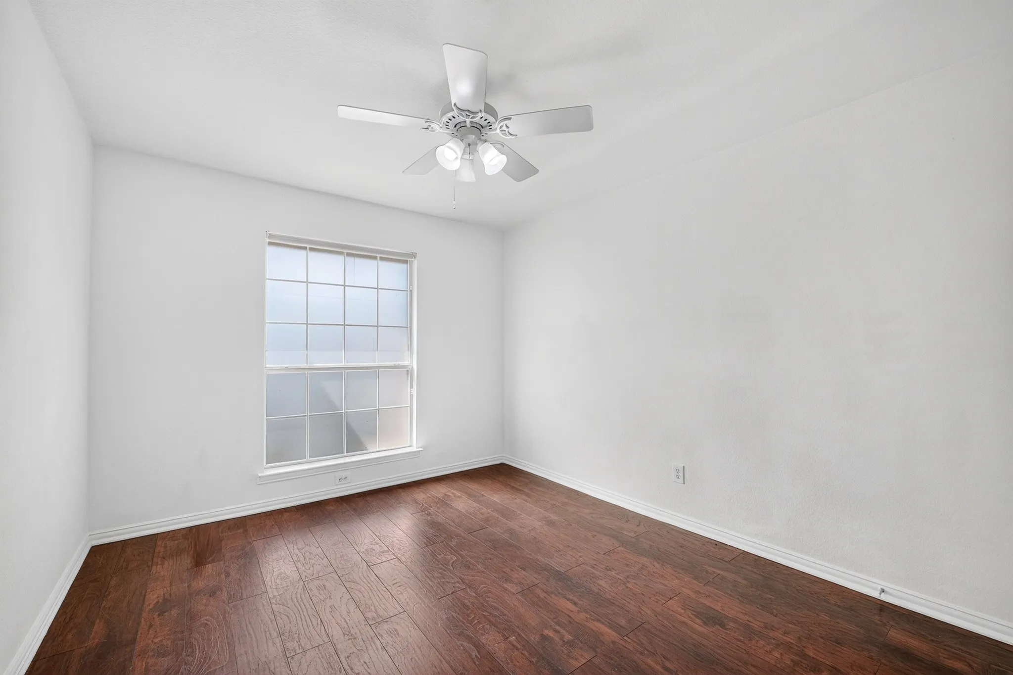 Neutral interior space featuring wood-finish flooring, a multi-pane window, and a central ceiling fan with integrated lighting