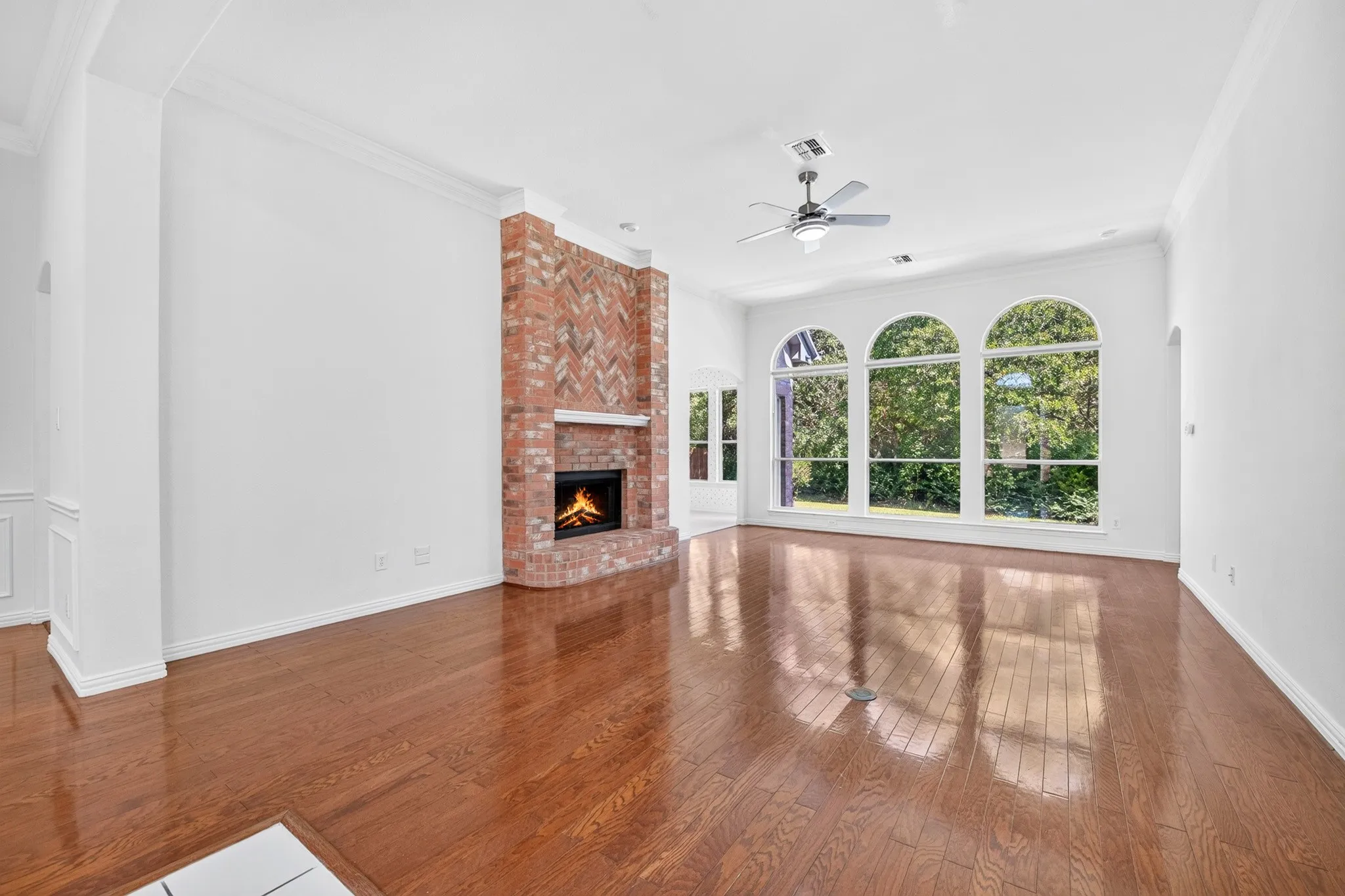 Living area featuring wood-finish flooring, a herringbone-patterned brick fireplace, three arched windows, and a ceiling fan