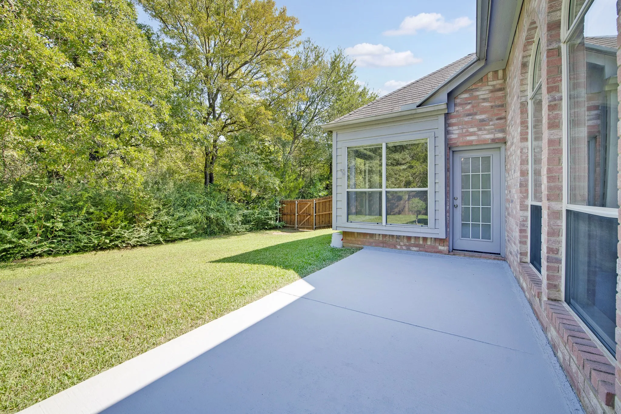 Concrete patio, brick exterior wall, multi-pane glass door, and expansive lawn featuring mature trees