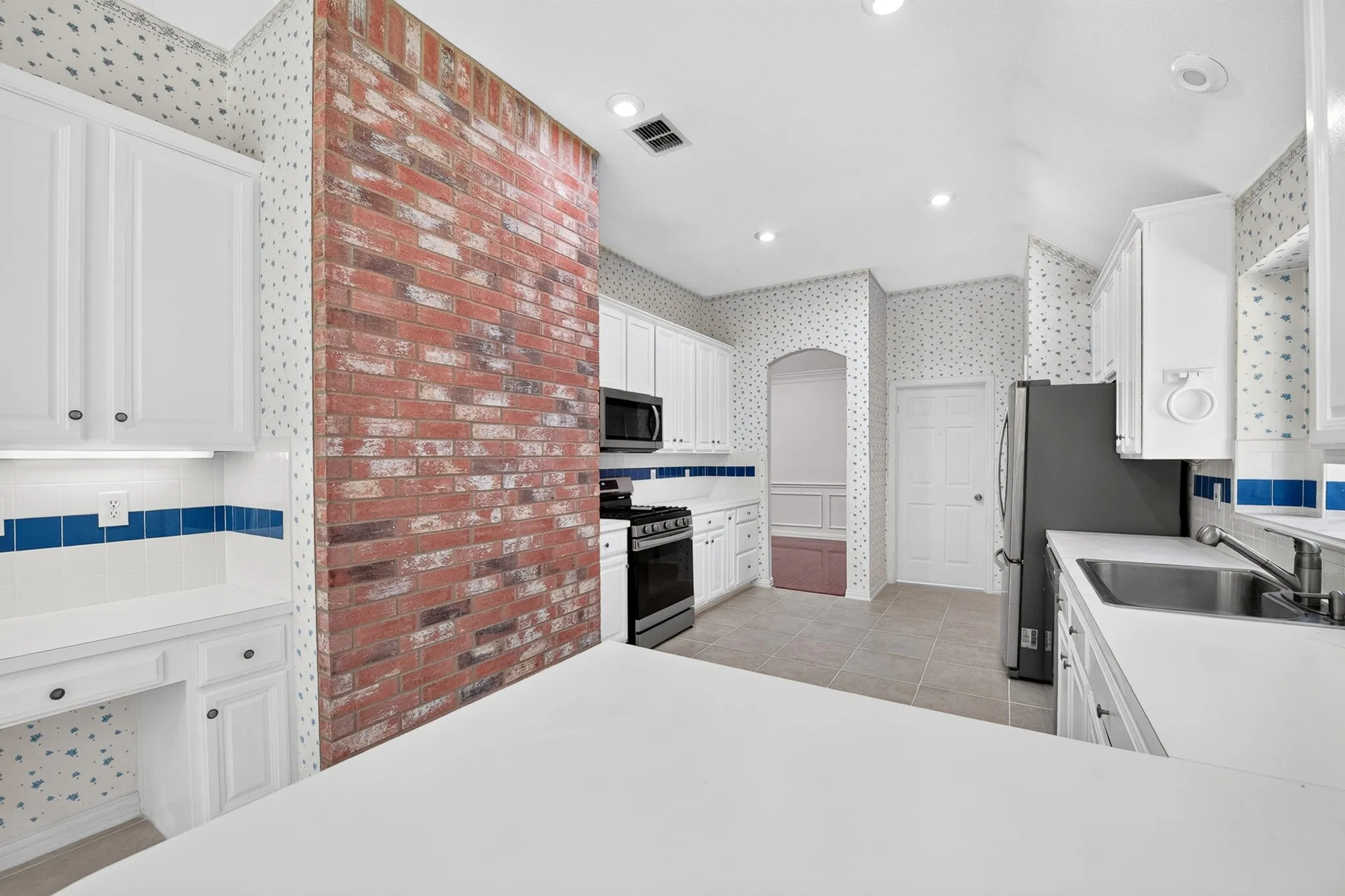 Kitchen featuring a floor-to-ceiling exposed brick accent wall, white cabinetry, solid surface countertops, stainless steel appliances, and tile flooring