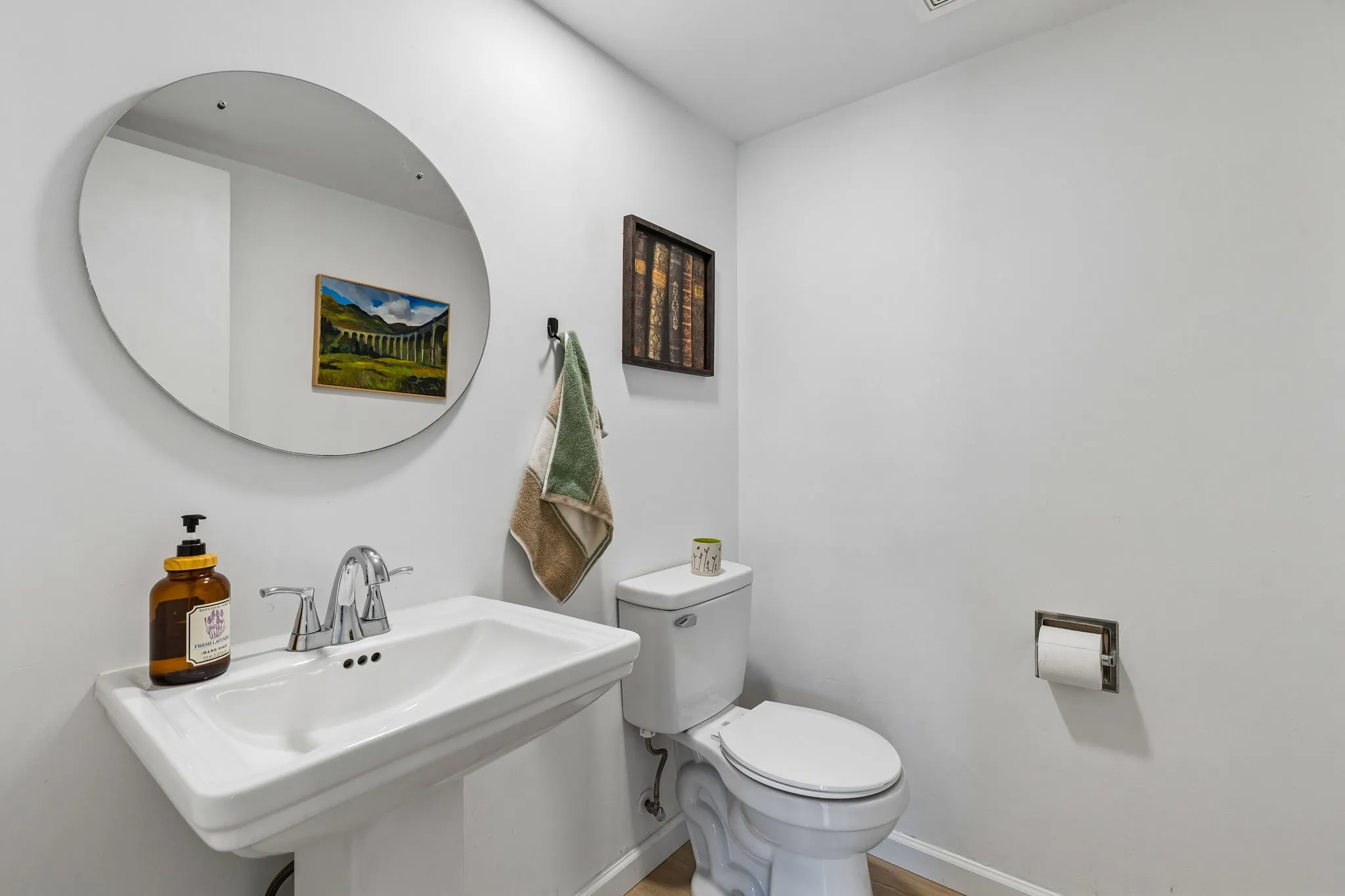 Pedestal sink with chrome dual-handle faucet, oval frameless wall mirror, and chrome toilet paper holder