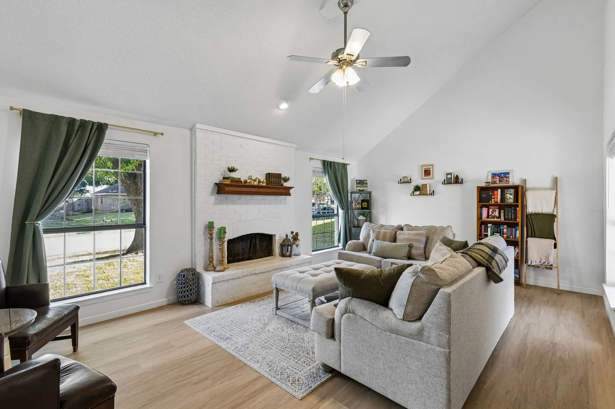 Vaulted ceiling, white-painted brick fireplace with a wood mantel, multi-pane windows, and light-toned wood-finish flooring