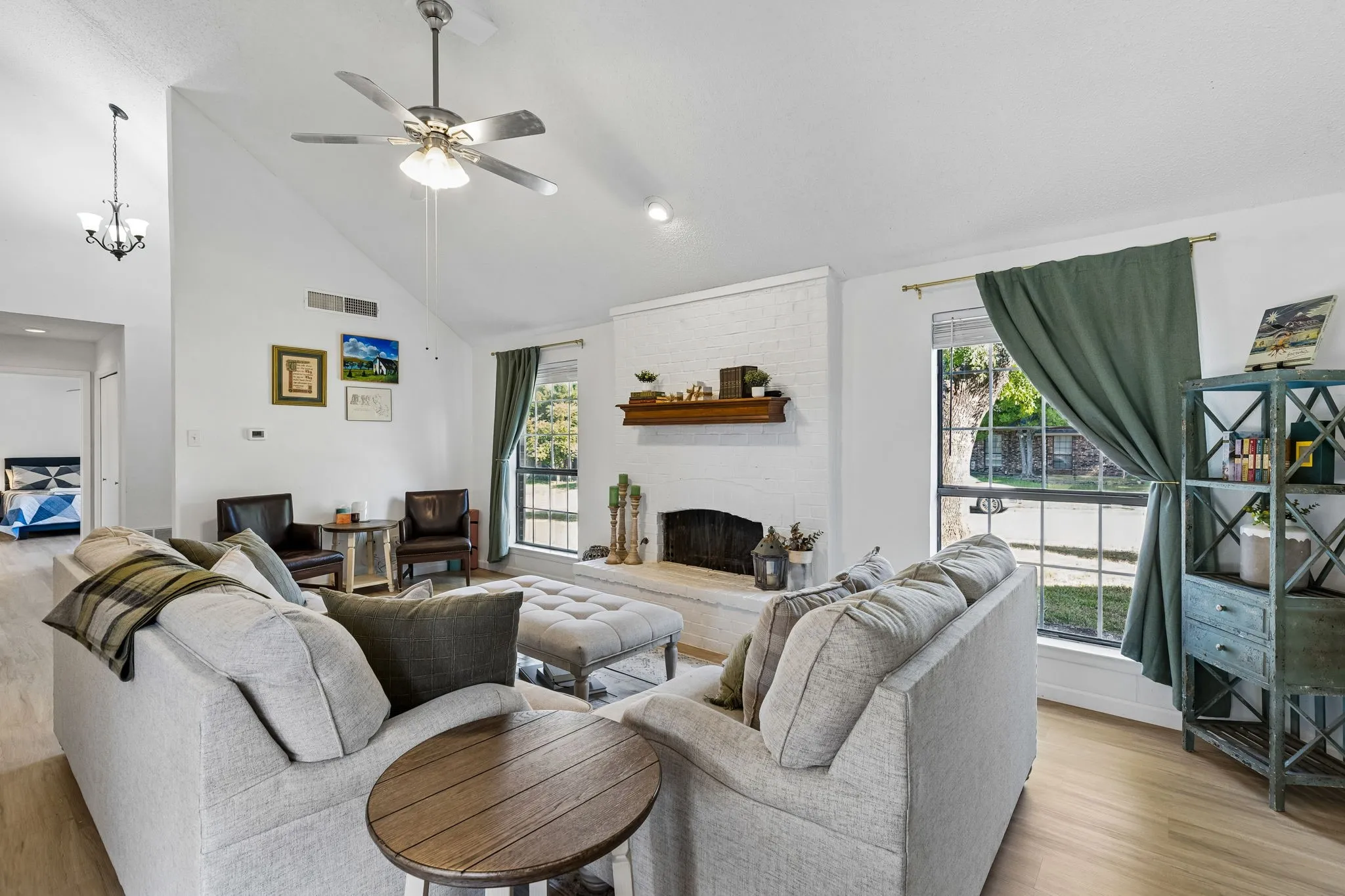 Living area featuring a vaulted ceiling, painted brick fireplace with a wood mantel, wood-finish flooring, ceiling fan, and large windows