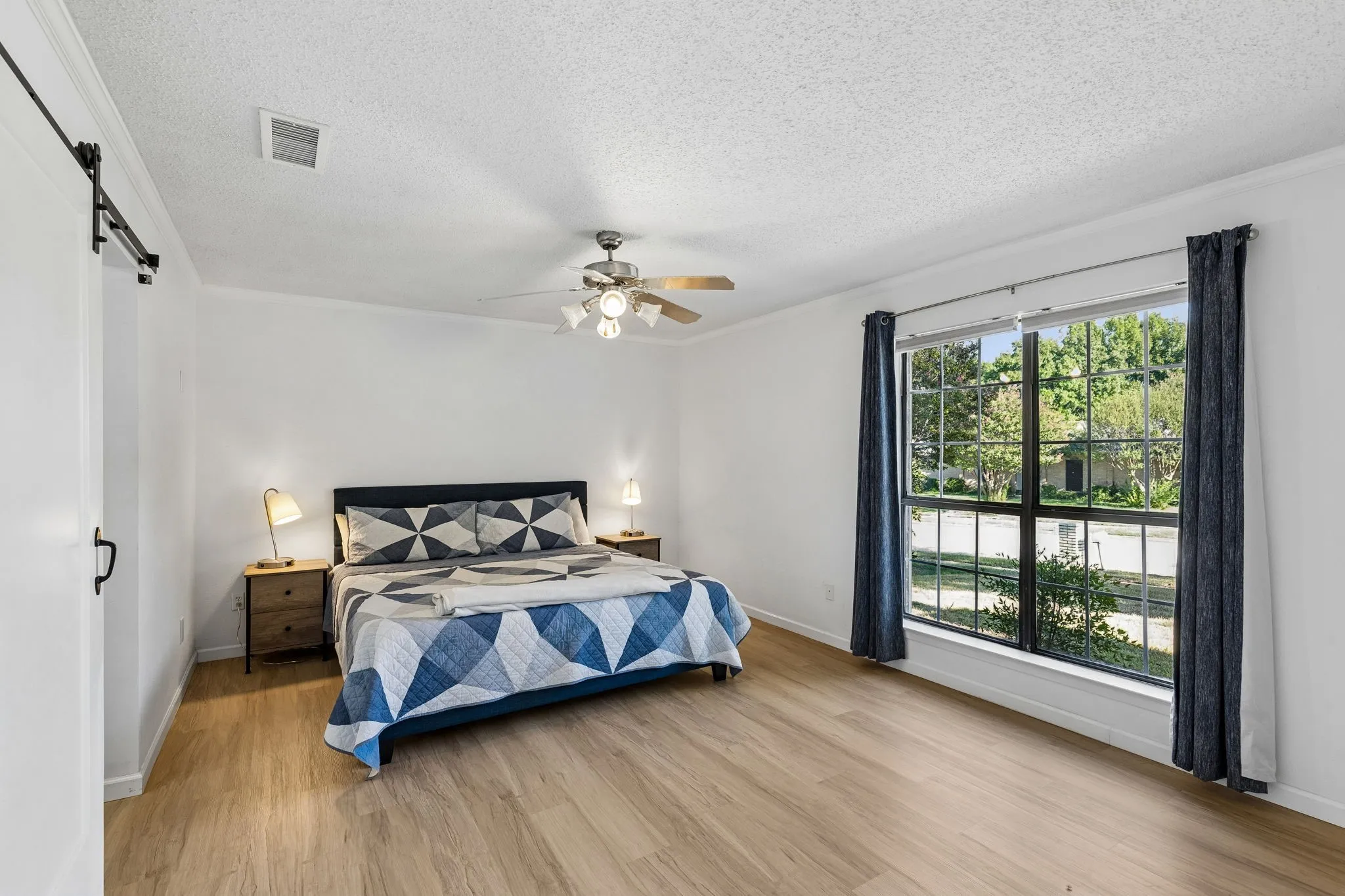 Bedroom featuring wood-finish flooring, a sliding barn door, a large multi-pane window, and a ceiling fan with integrated lighting