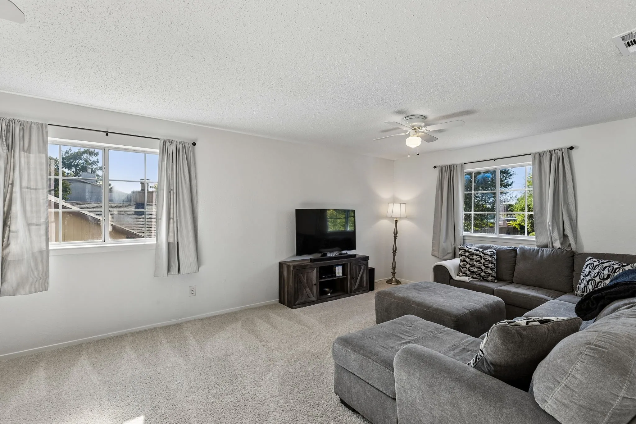 Neutral-toned interior space featuring light-colored wall-to-wall carpeting, dual windows with multi-pane grids, and a ceiling-mounted light fixture with a multi-blade fan