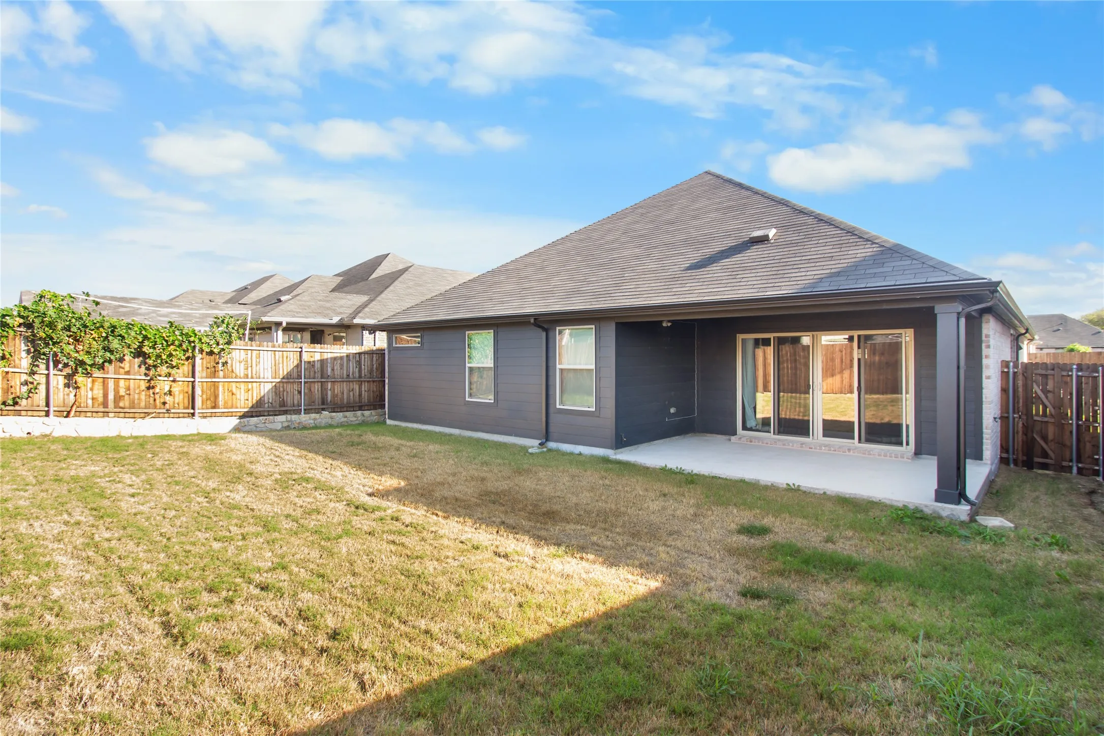 Rear exterior featuring dark-toned siding, a covered concrete patio, sliding glass door access, and a fenced yard with wood perimeter fencing