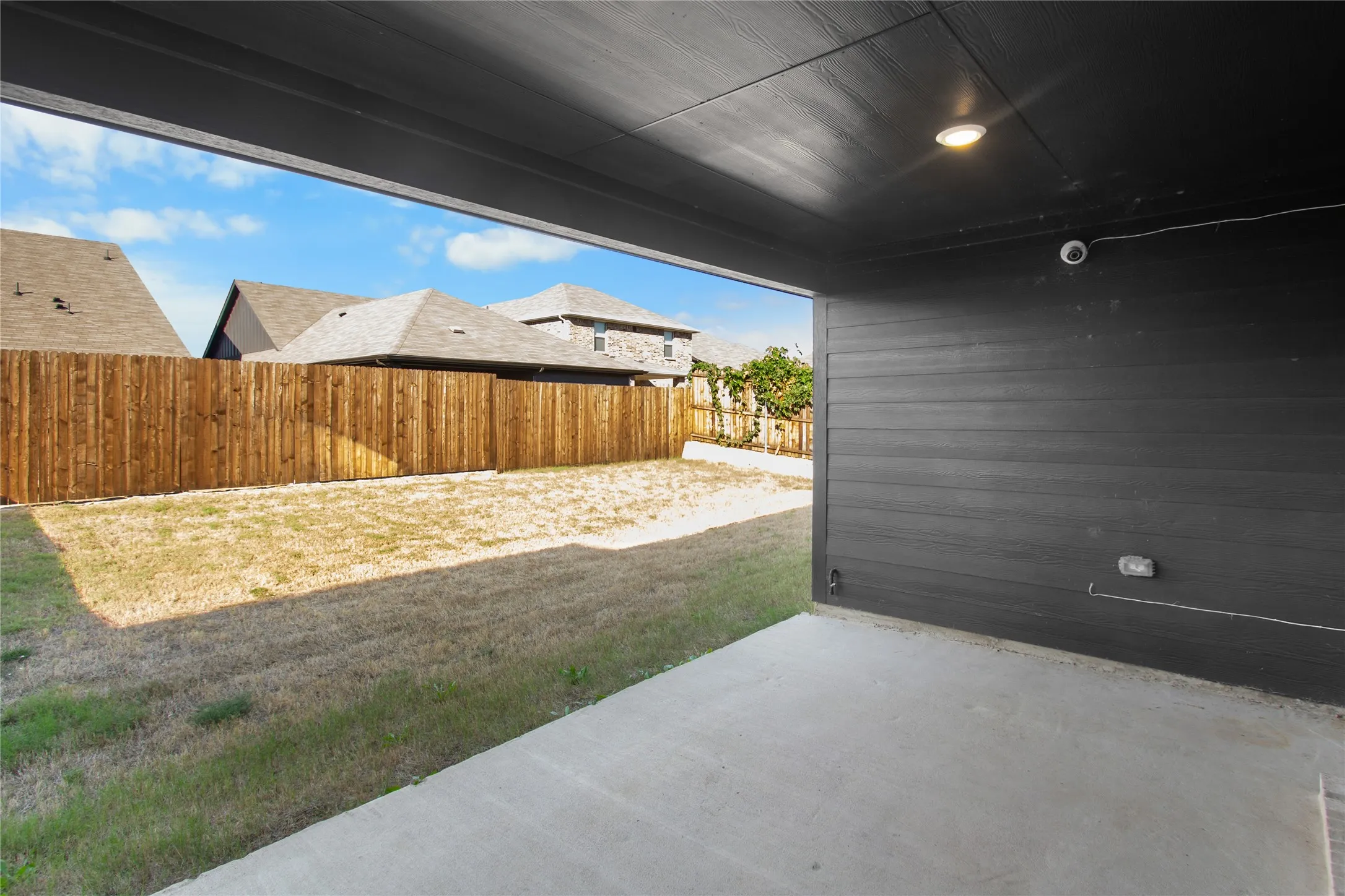 Covered concrete patio featuring dark horizontal siding, recessed lighting, and an enclosed backyard with wood privacy fencing