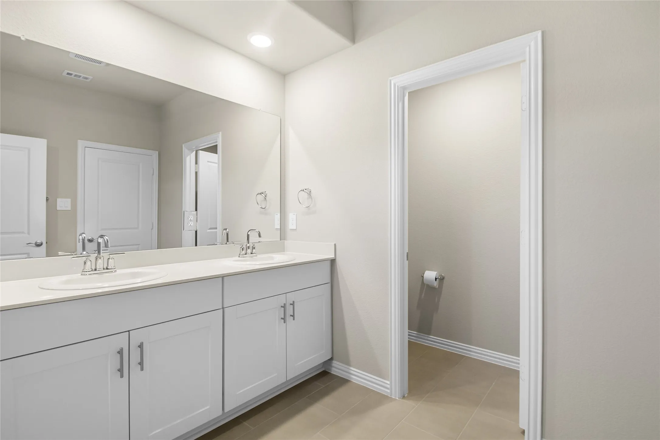 Dual-sink vanity featuring white cabinetry, solid-surface countertop, chrome fixtures, and a wall-to-wall mirror with recessed ceiling lighting