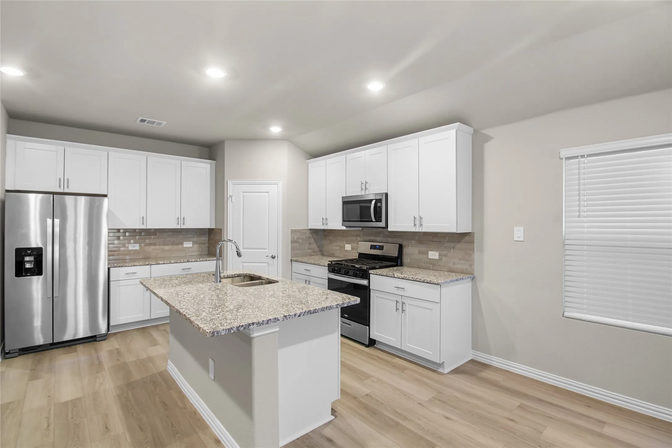 Kitchen featuring white shaker-style cabinetry, stone-surface countertops, wood-finish flooring, a central island with an undermount sink, and stainless steel appliances