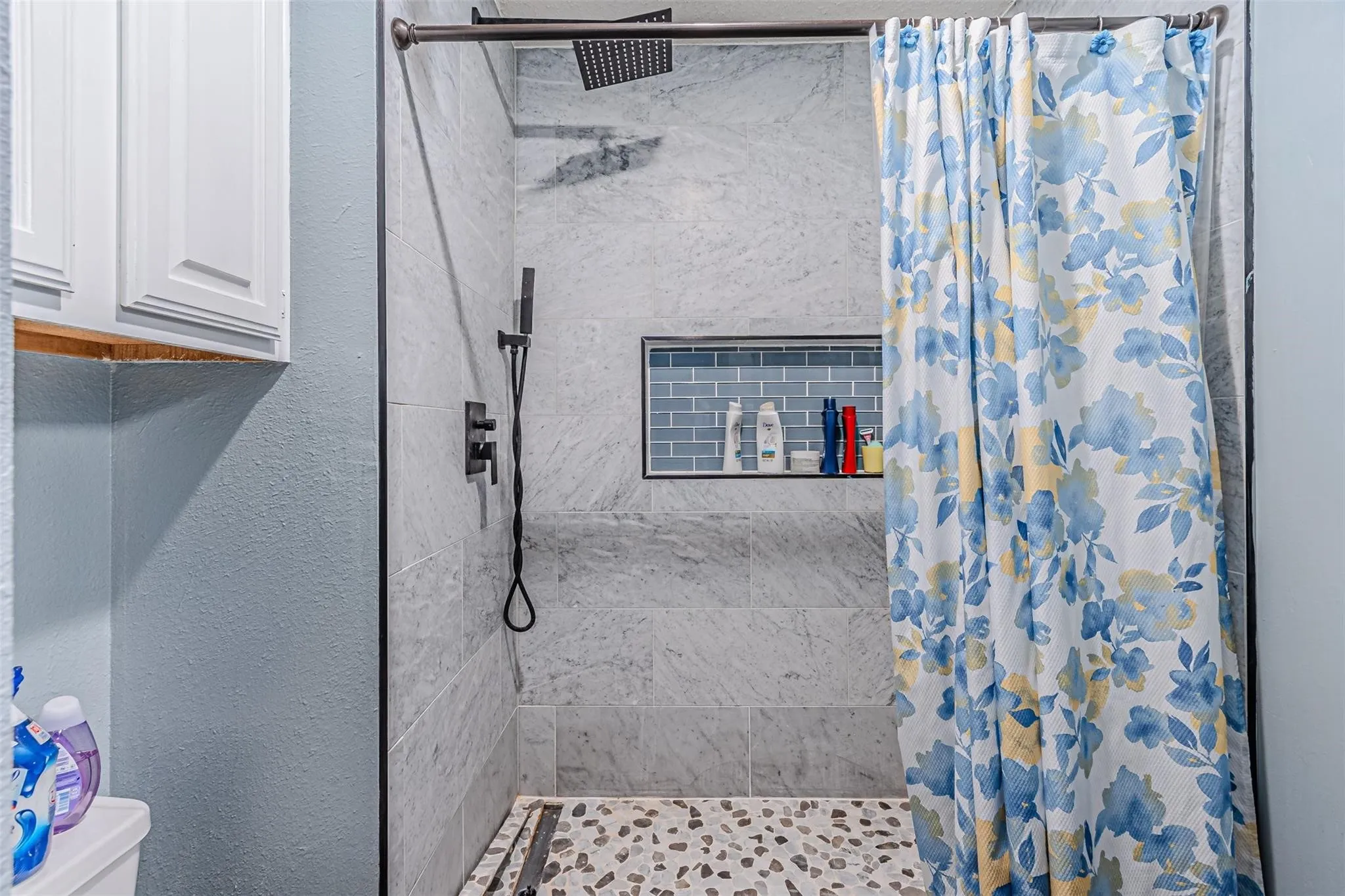 Walk-in shower featuring large-format stone-tile surround, blue subway-tile recessed niche, rainfall showerhead with matte black fixtures, and decorative pebble-stone flooring