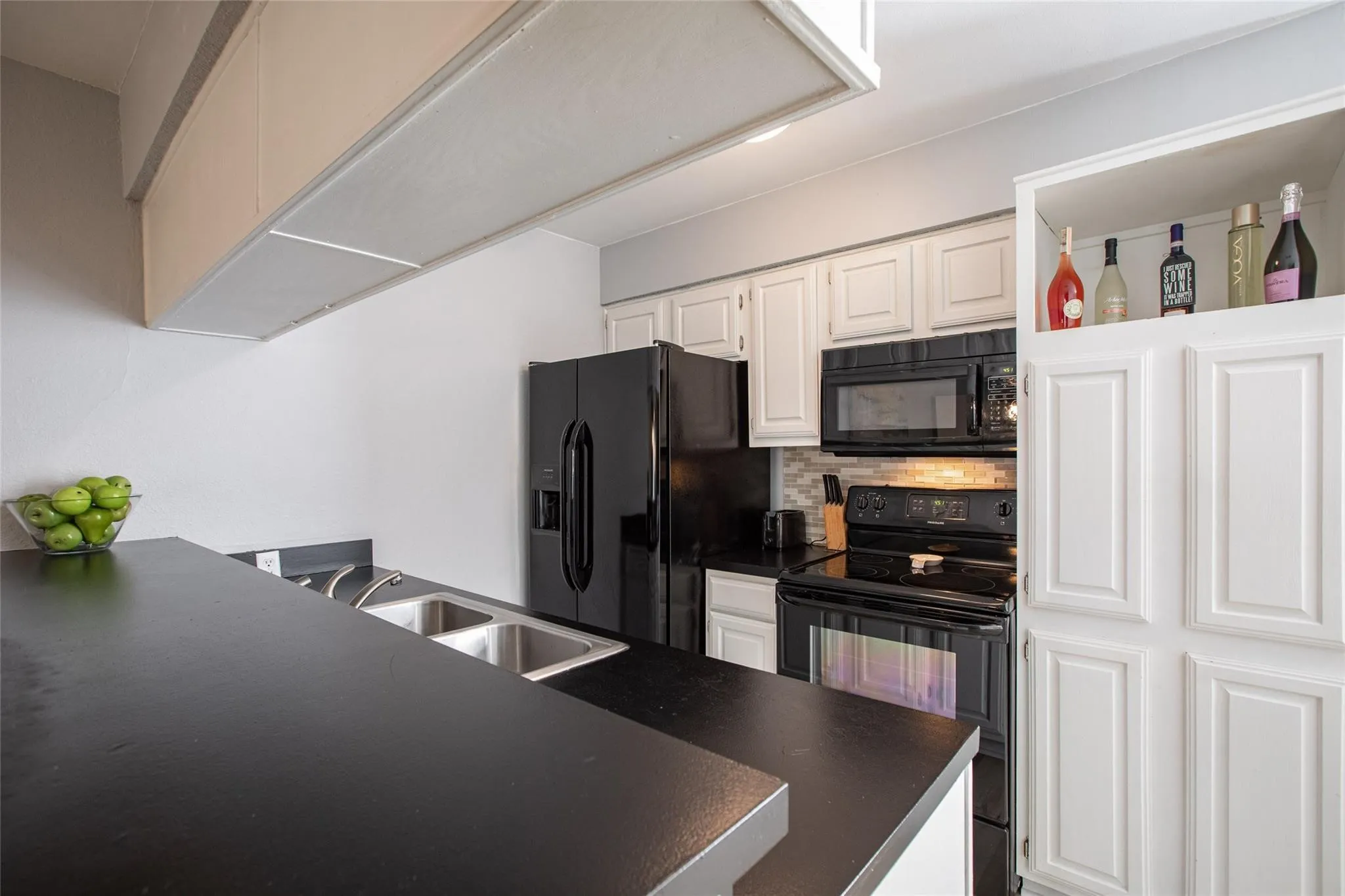 Kitchen featuring black countertops, white shaker-style cabinetry, a built-in pantry with open shelving, a double-basin stainless steel sink, and black appliances
