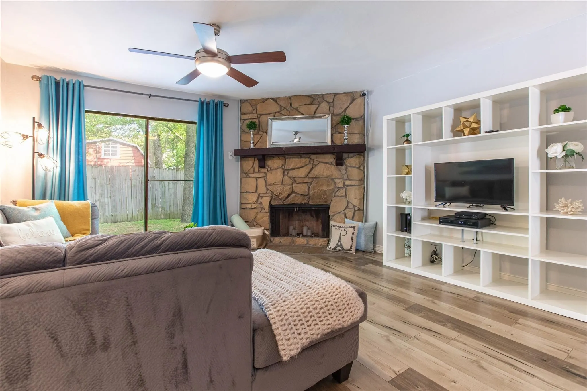 Floor-to-ceiling stone fireplace with a wooden mantel, wood-finish flooring, built-in white shelving unit, and a ceiling fan