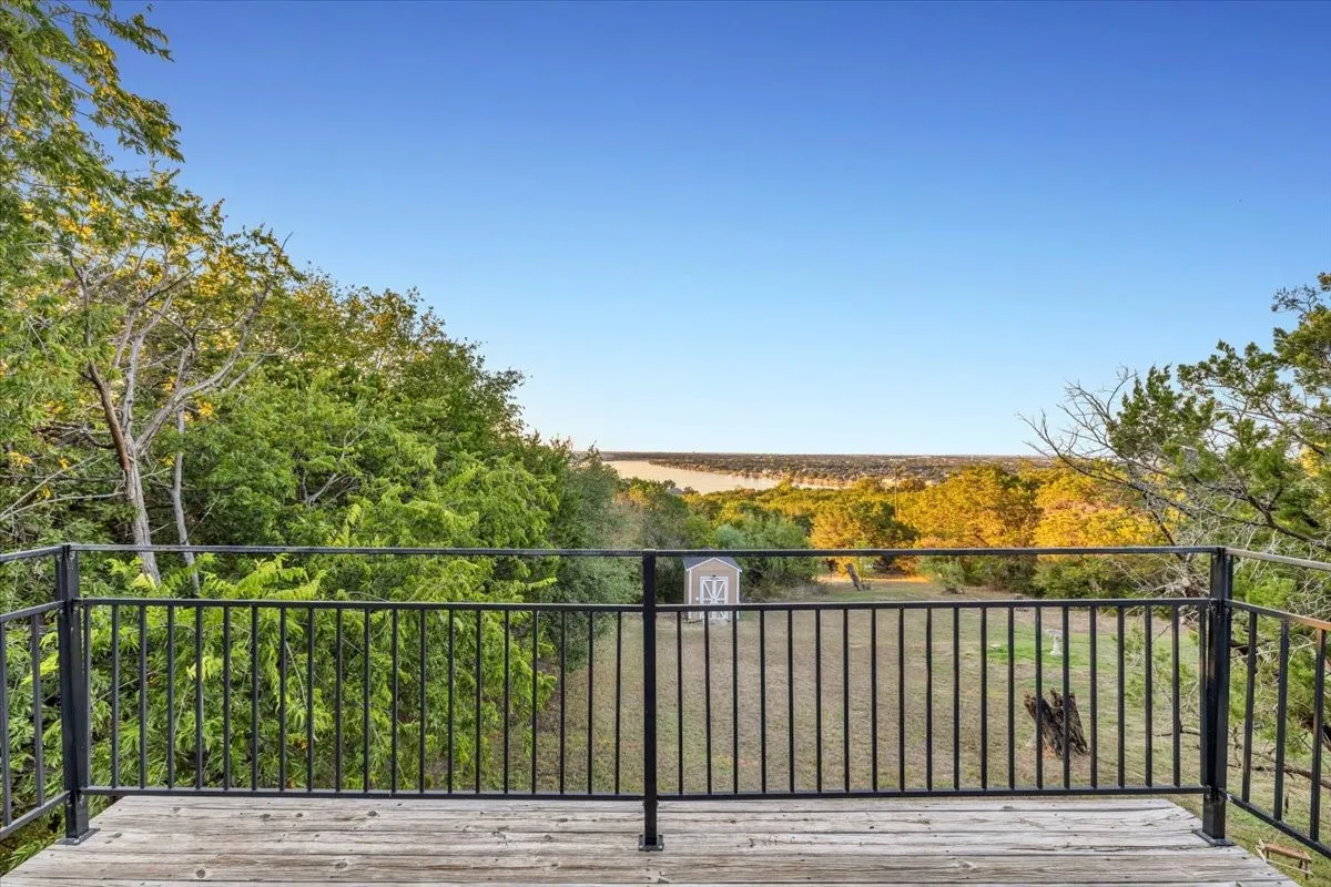 Elevated wooden deck with black metal railing, overlooking a fenced outdoor area featuring mature trees and a detached storage structure